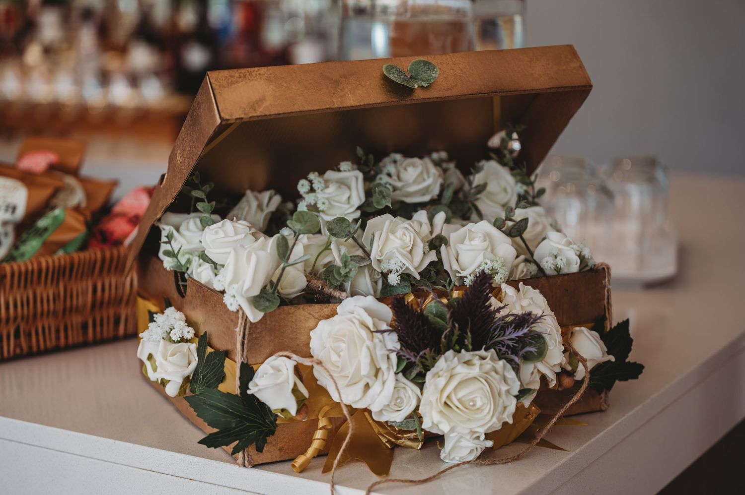 White roses and greenery arranged inside a rustic brown wooden box displayed on a white surface.