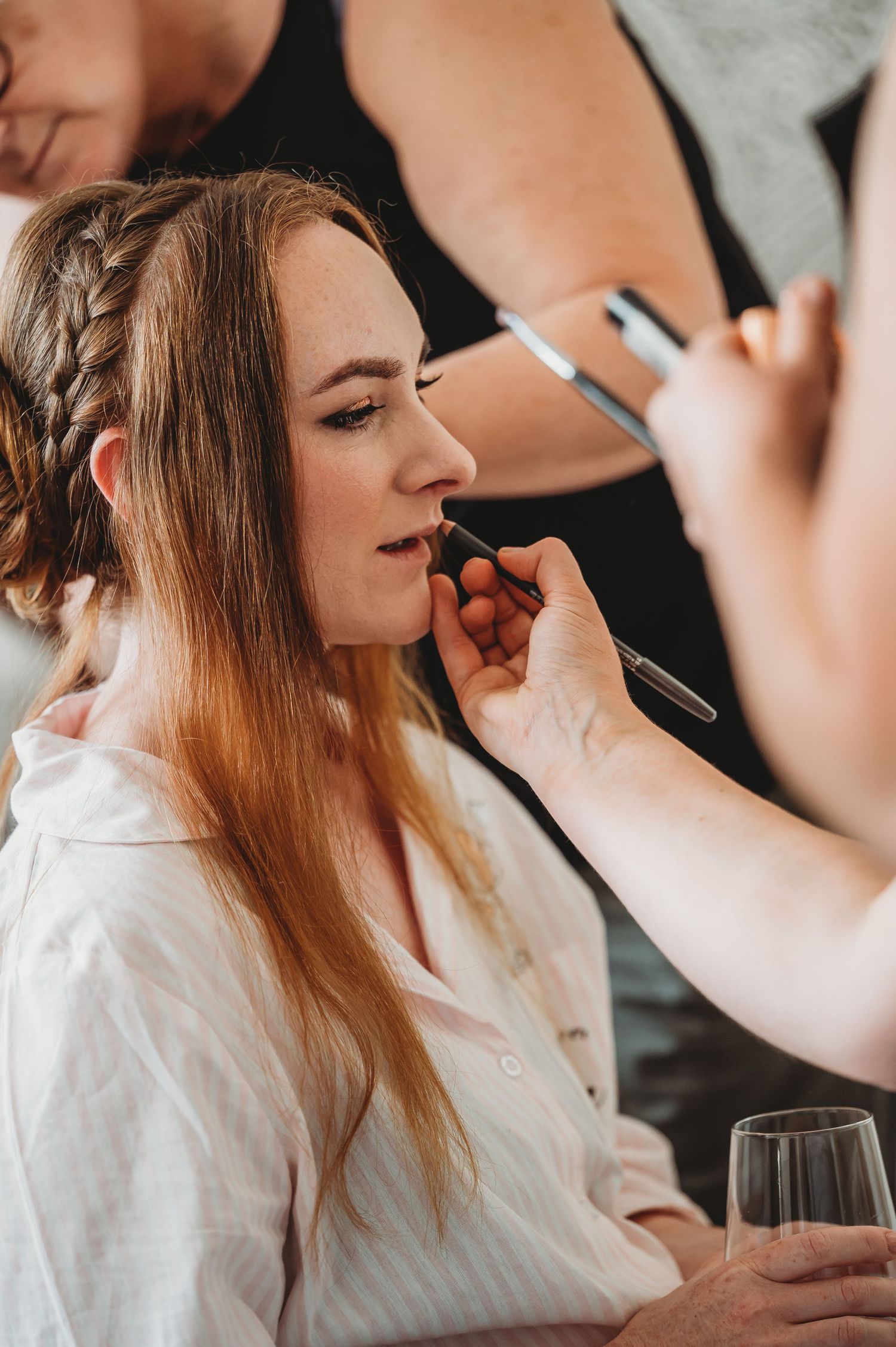 Makeup artist applies cosmetics to bride during wedding preparation while client holds champagne glass.