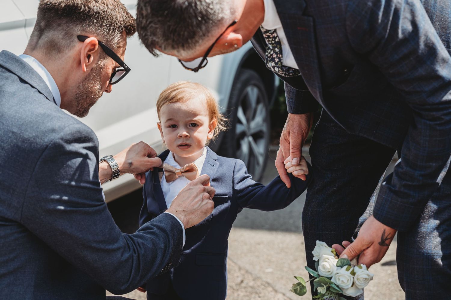Two men in formal attire adjust a young boy's bowtie during what appears to be a wedding preparation.