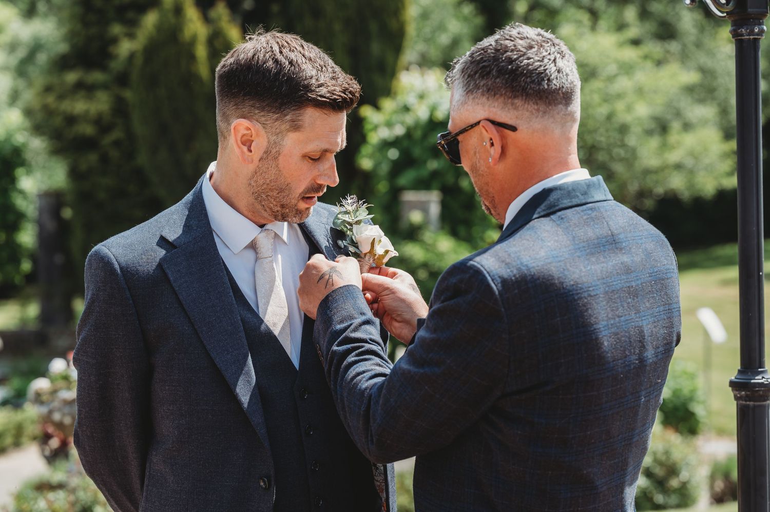 Two men in gray suits stand close together while adjusting a boutonniere at an outdoor event.