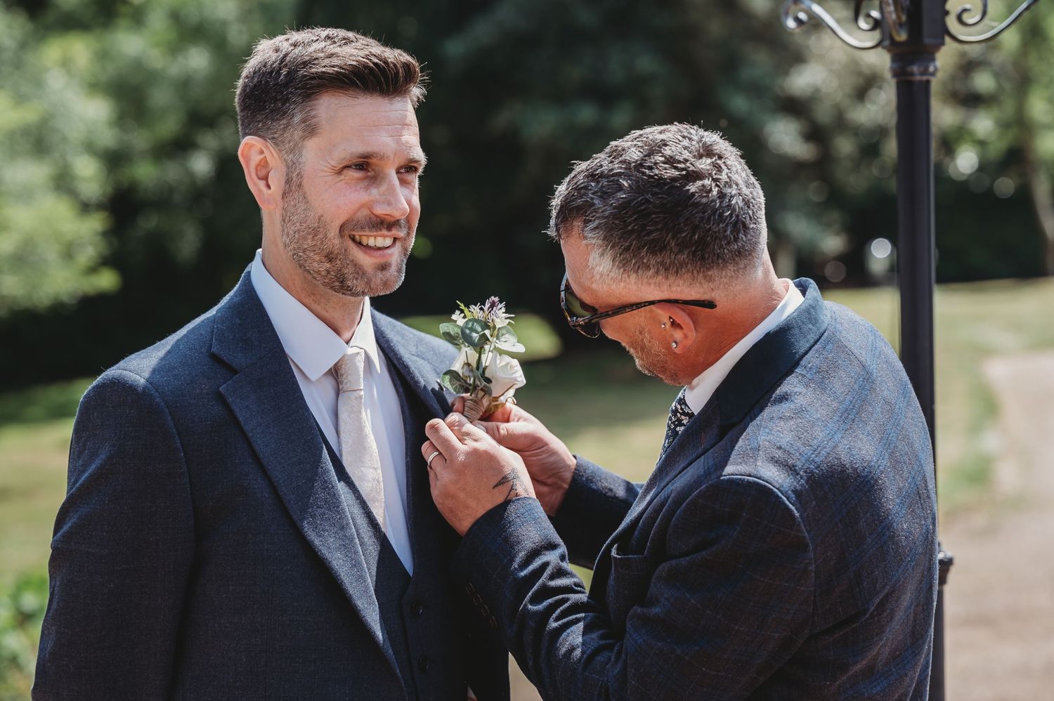 Two people in navy suits share a joyful moment during an outdoor wedding ceremony as one adjusts the other's boutonniere.