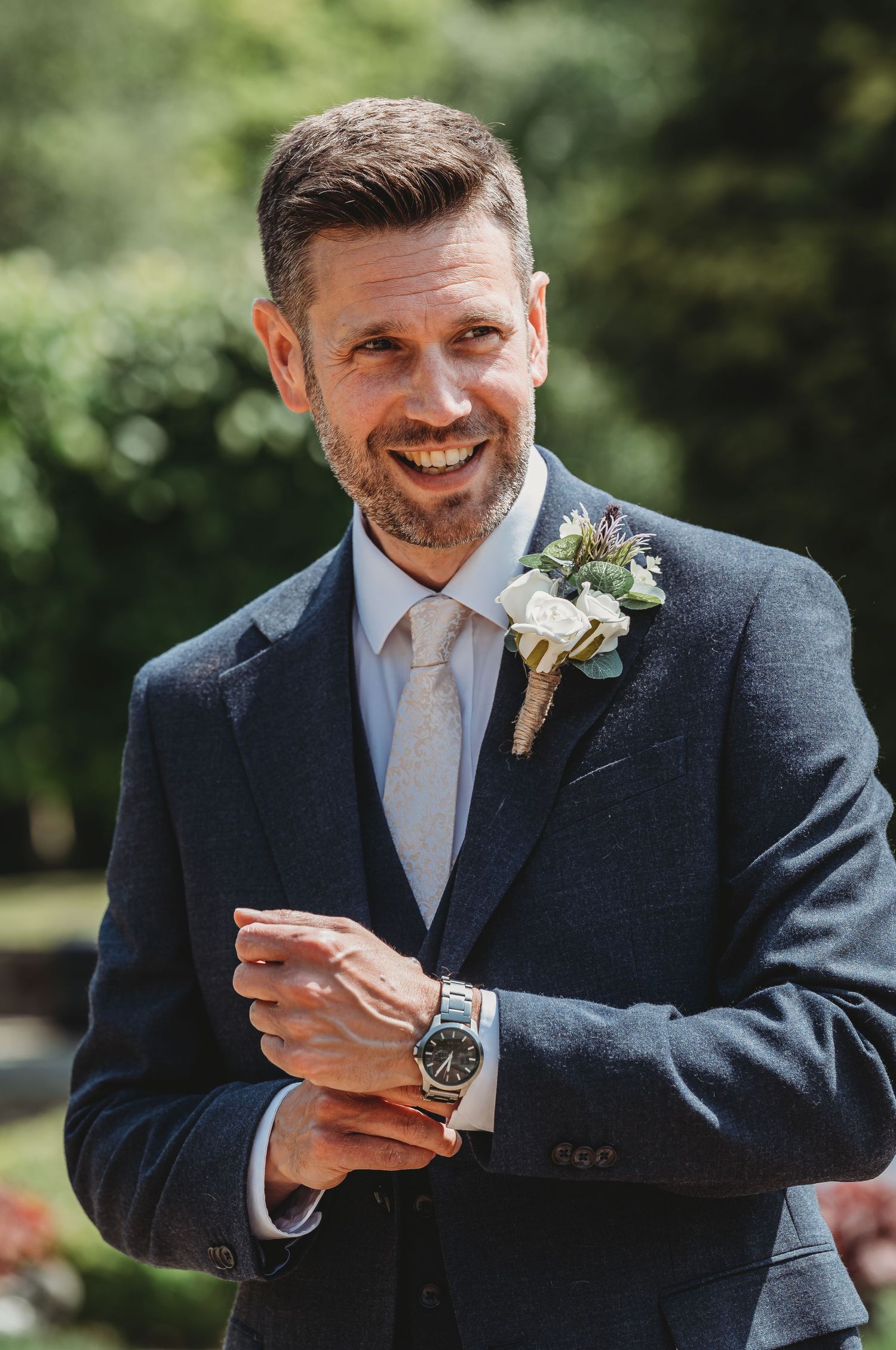 A groom in a navy suit with boutonniere smiles while adjusting his watch on a sunny wedding day outdoors.