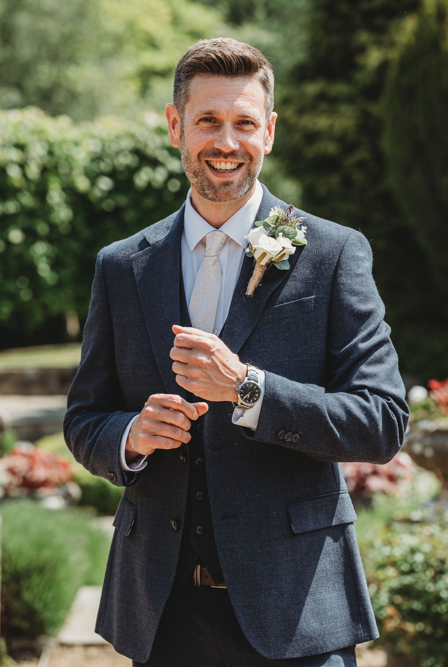A groom wearing a navy suit and light blue tie smiles while adjusting his boutonniere in an outdoor garden setting.