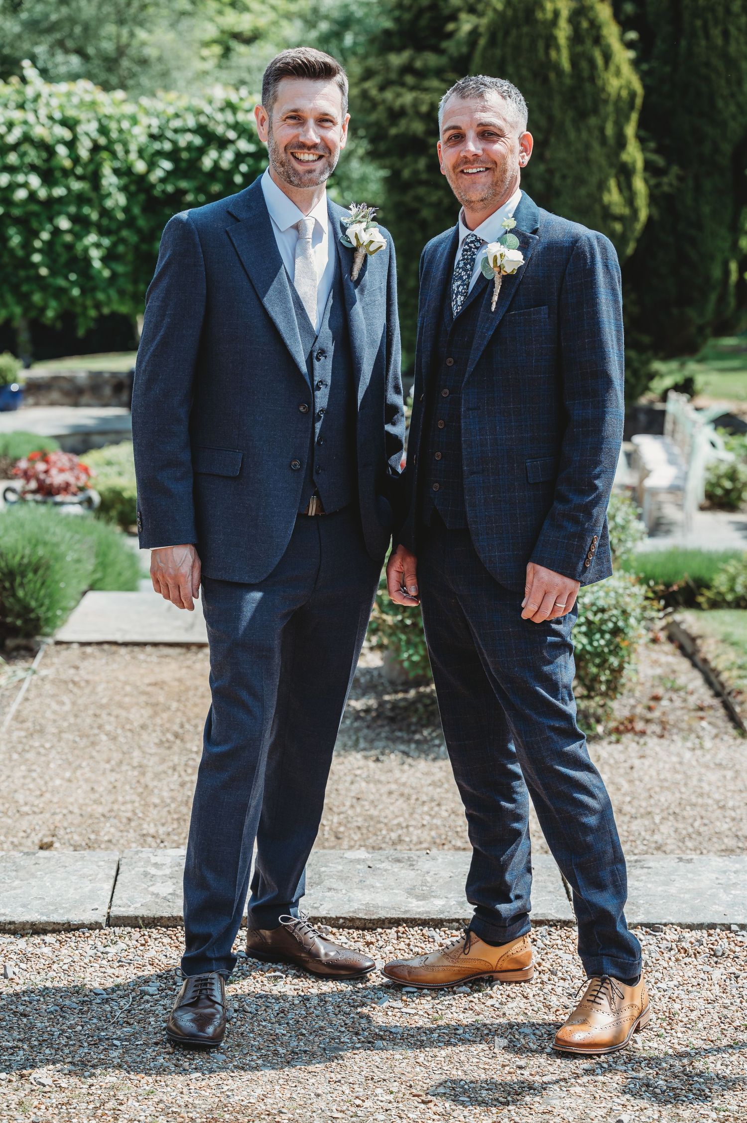 Two men in navy blue suits and boutonnieres stand on a garden path with greenery and flowers in the background.