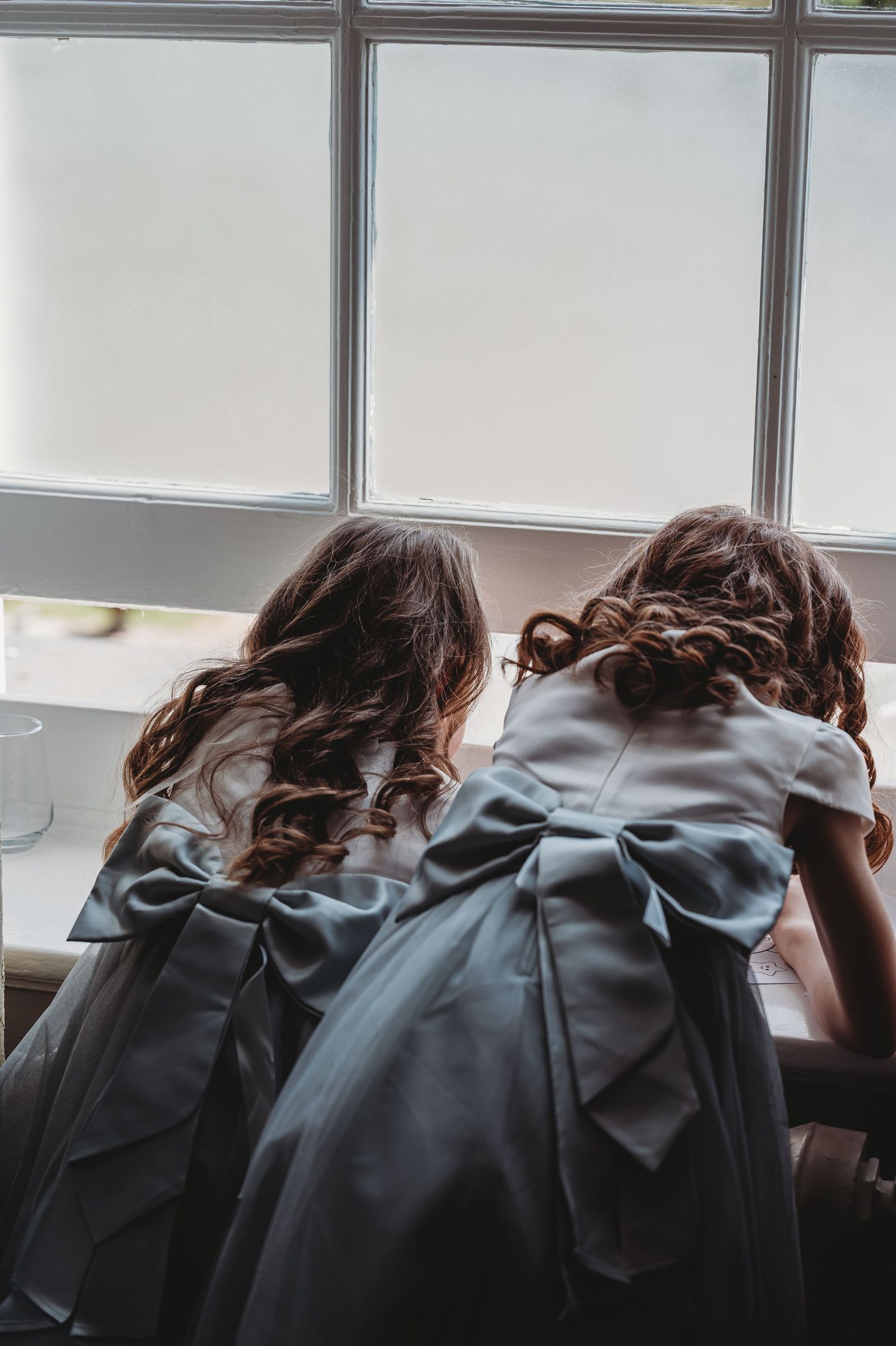 Two bridesmaids in matching blue dresses with bows look out a window during wedding preparations.