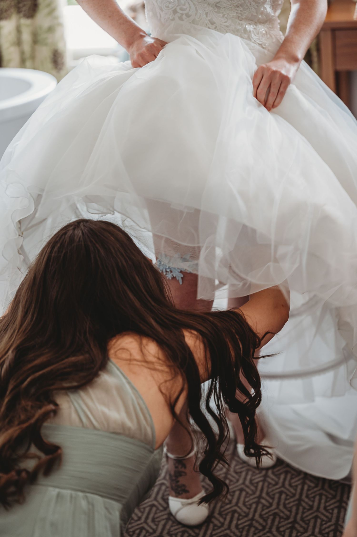 Helper adjusts the white lace garter under a flowing wedding dress while crouching on patterned carpet.