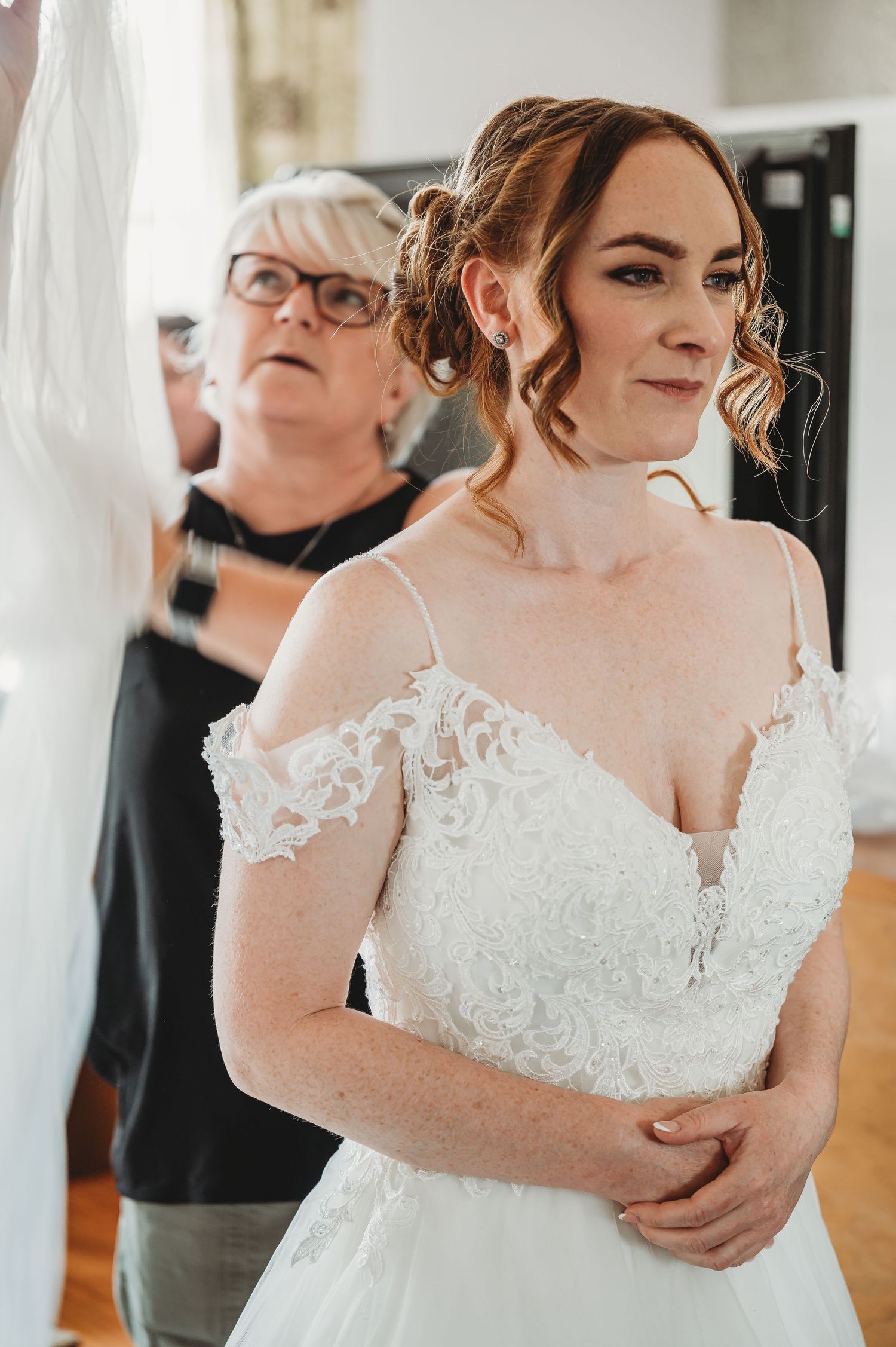A bride wearing an elegant white lace off-shoulder wedding gown gets final adjustments in a sunlit room.