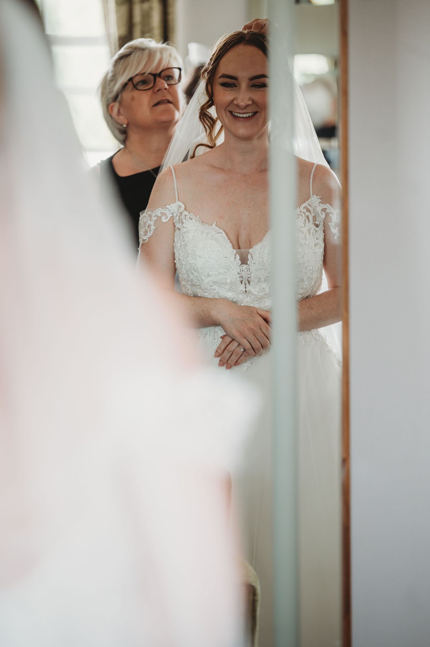 Candid bridal preparation moment captured through doorway showing elegant lace wedding dress and joyful expressions.