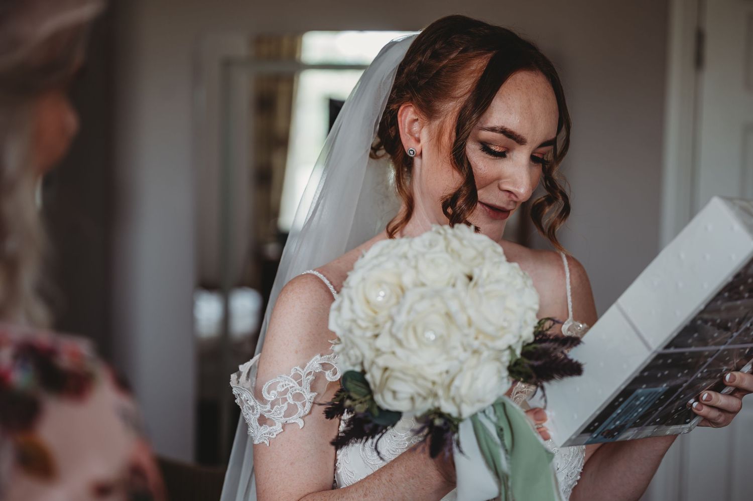 A bride in white lace reads while holding a classic white rose bouquet during wedding preparations.