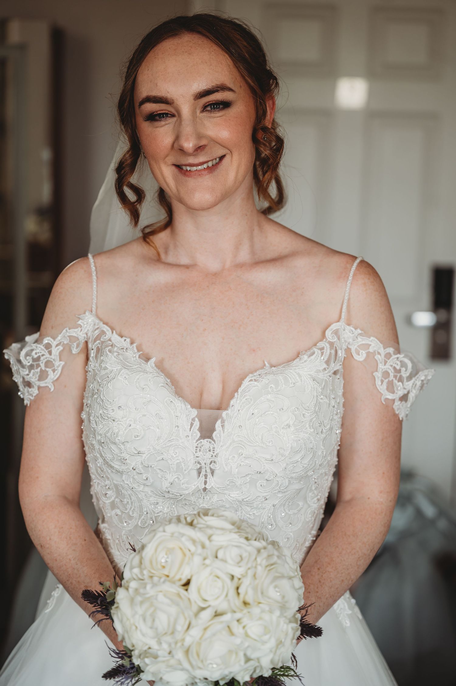 A bride in a delicate lace wedding dress with cold shoulder details smiles while holding a white rose bouquet.