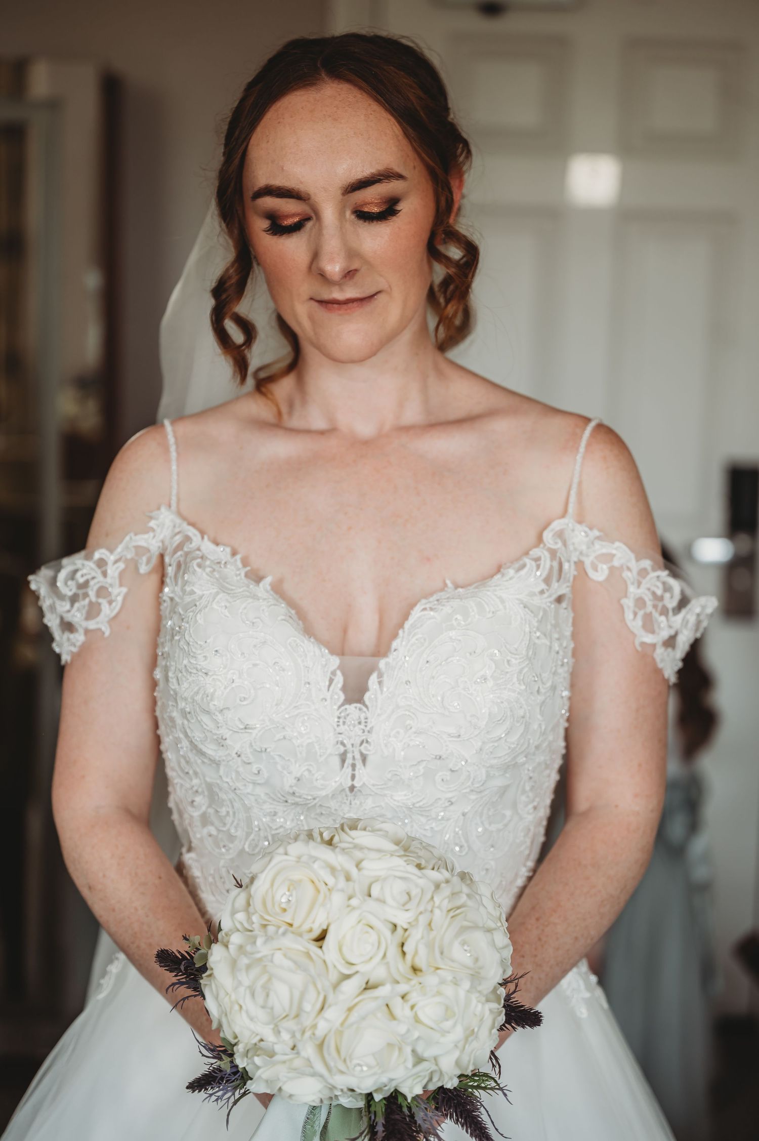 Bride in delicate off-shoulder lace wedding gown holds elegant white rose bouquet while looking down with soft makeup.