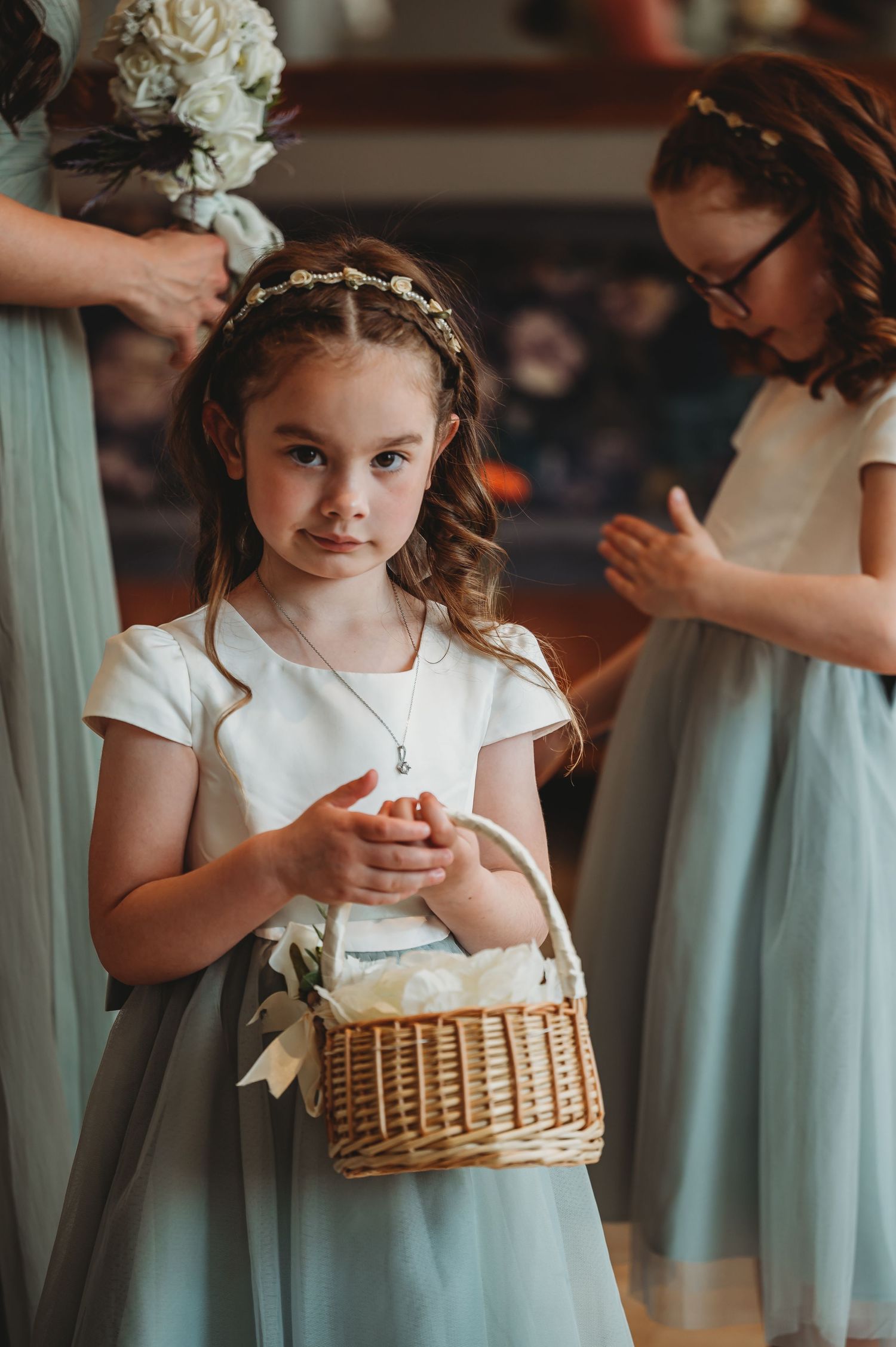 Young flower girl in white and mint dress holds wicker basket at wedding ceremony preparations.