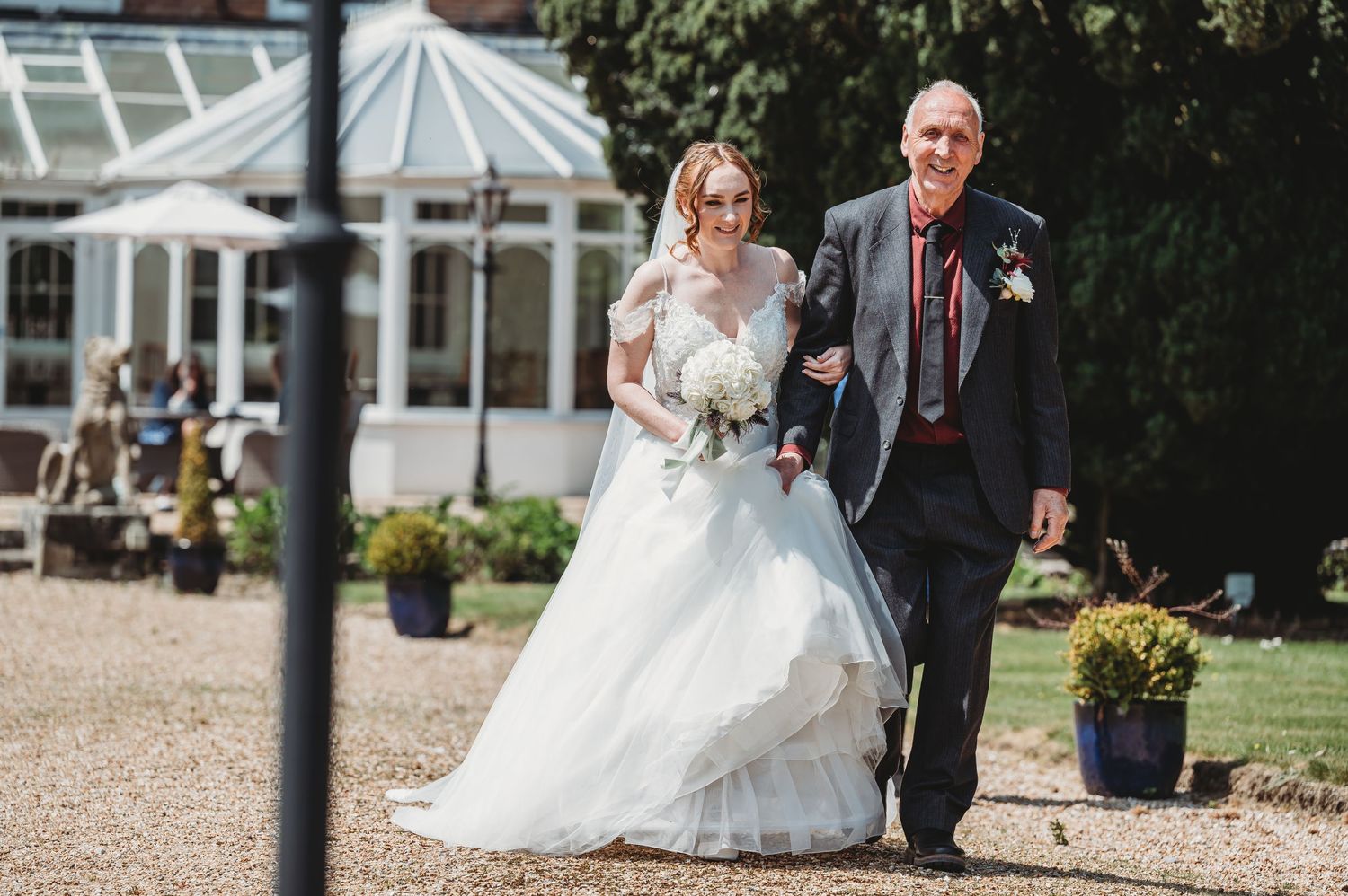 A wedding couple walks along a gravel path outside a conservatory with decorative potted plants on a sunny day.