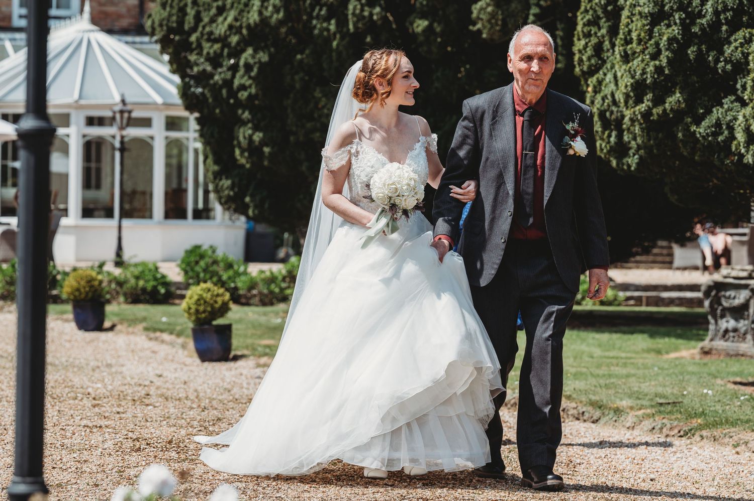A couple walks along a gravel path outside of an elegant venue with a conservatory in the background on their wedding day.