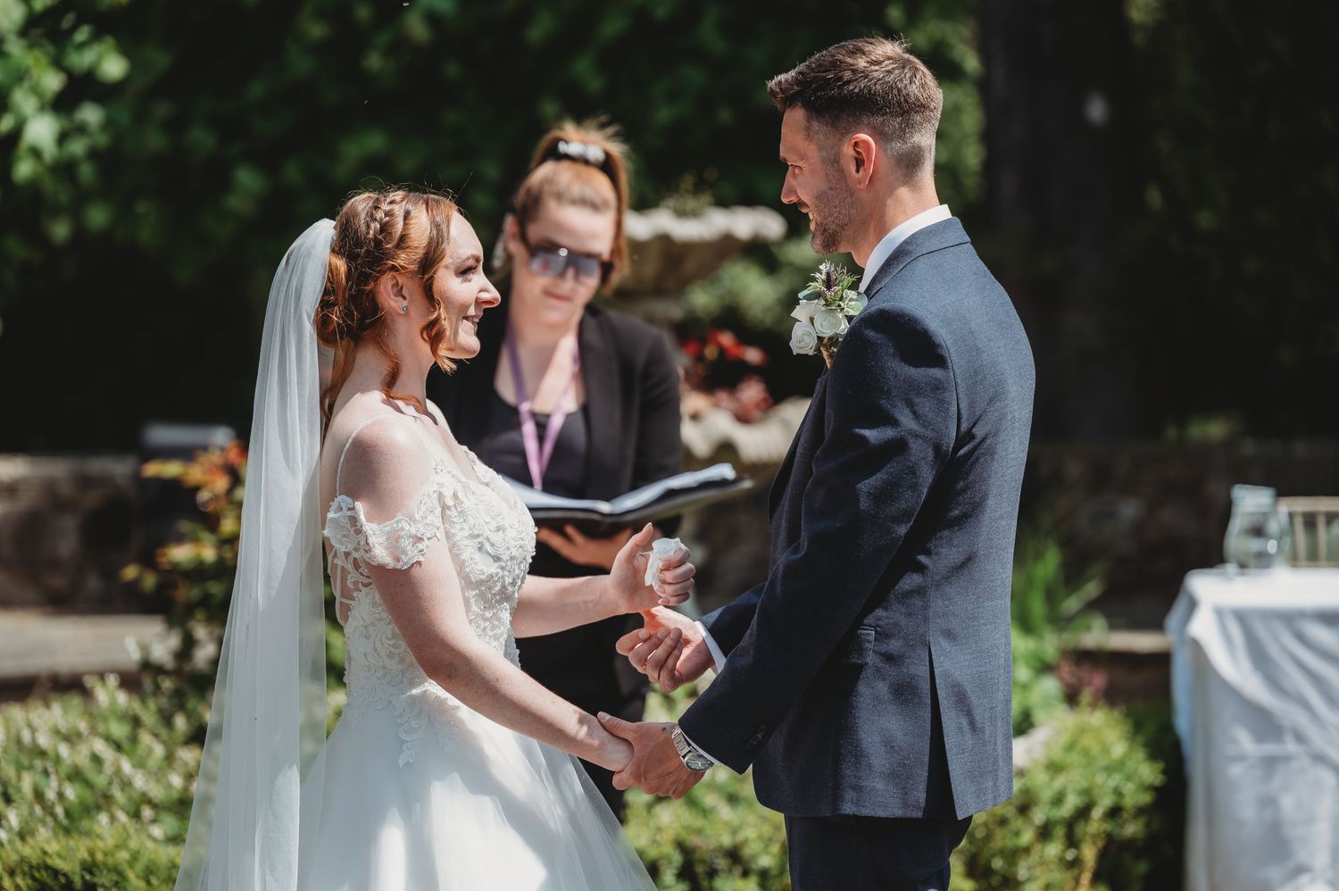 An outdoor wedding ceremony takes place in a lush garden setting with an officiating minister at the altar.