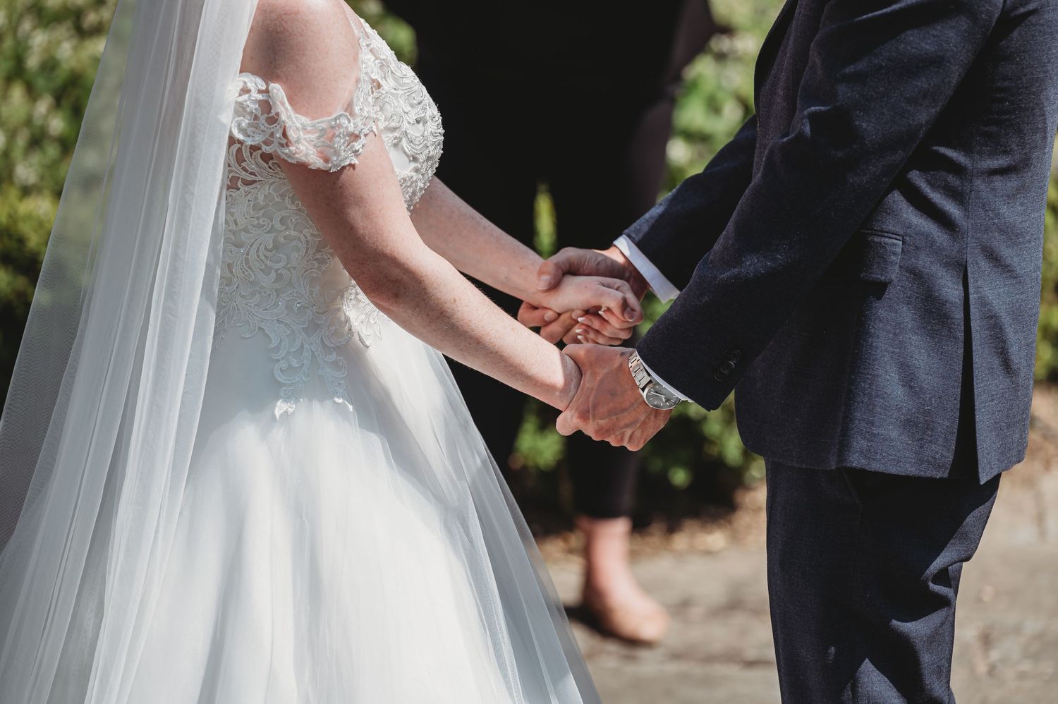 A close-up shot of clasped hands during a wedding ceremony, showing a white lace dress and navy suit.
