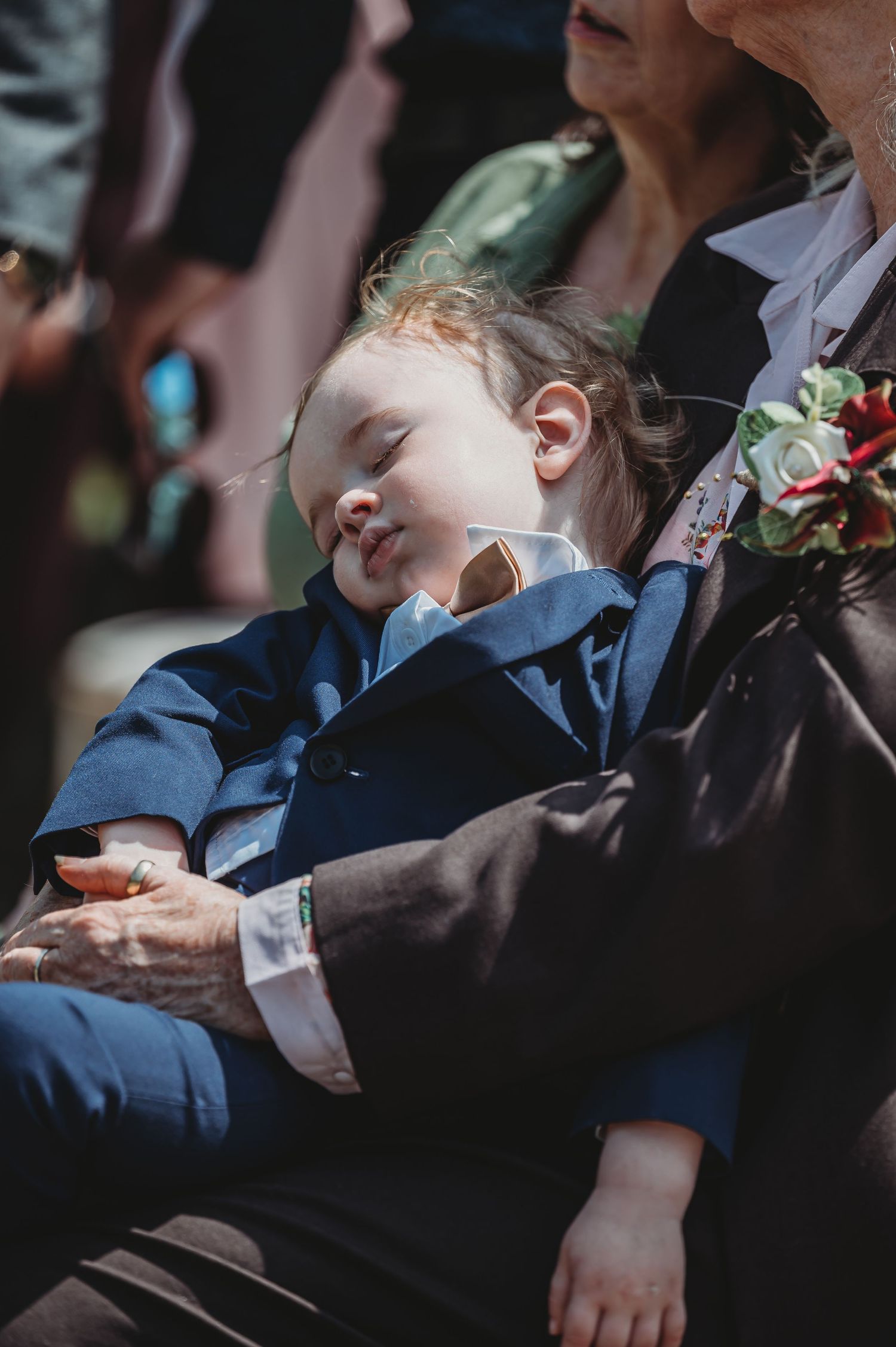 Young child in a blue suit napping during a formal event while being held, with flowers visible in background.
