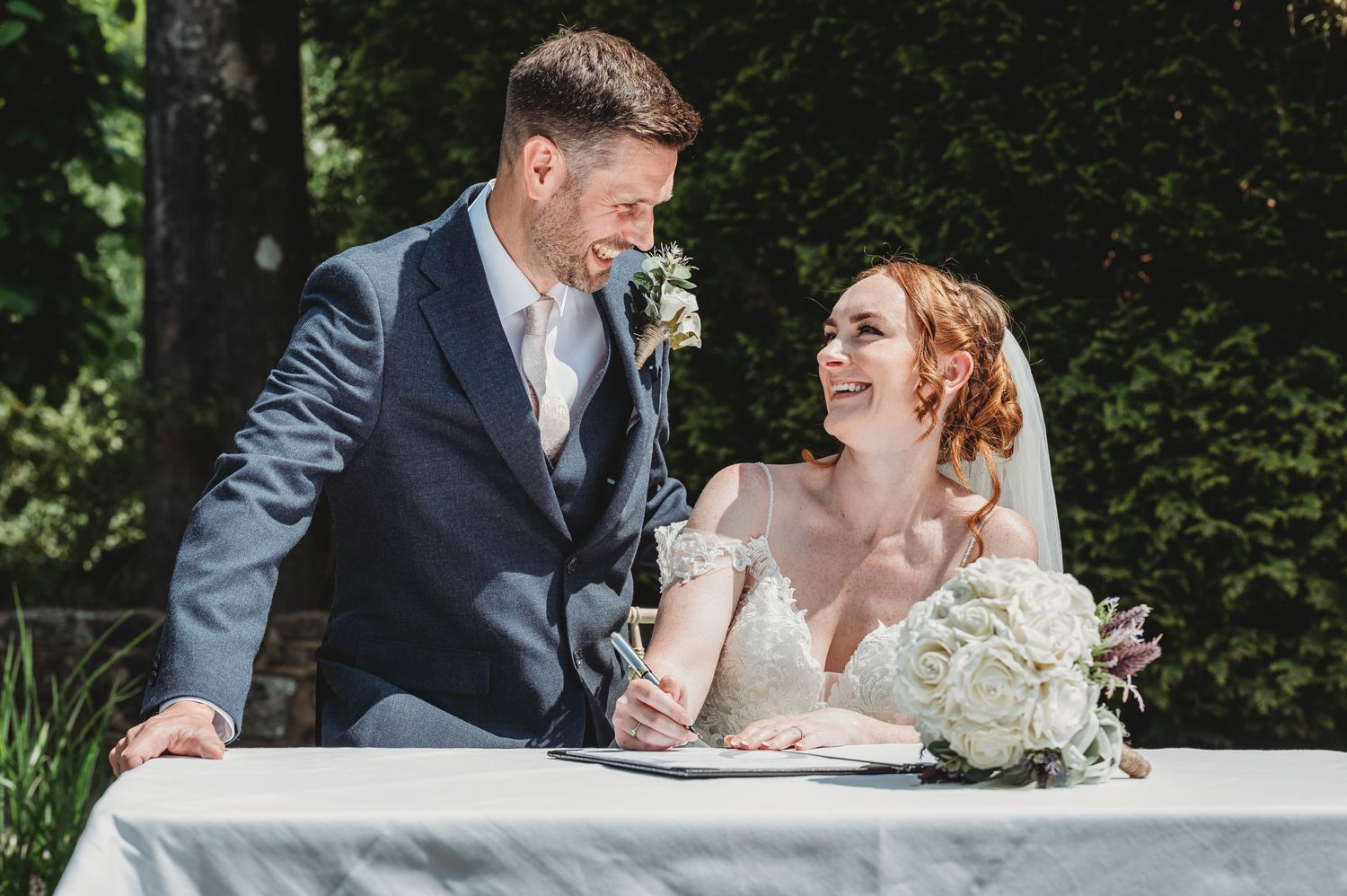 Wedding couple shares joyful laughter while sitting at a table outdoors with flowers and champagne.
