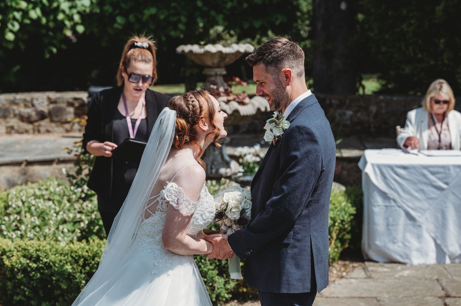 A couple shares their wedding vows during an outdoor garden ceremony with an officiant and guests present.