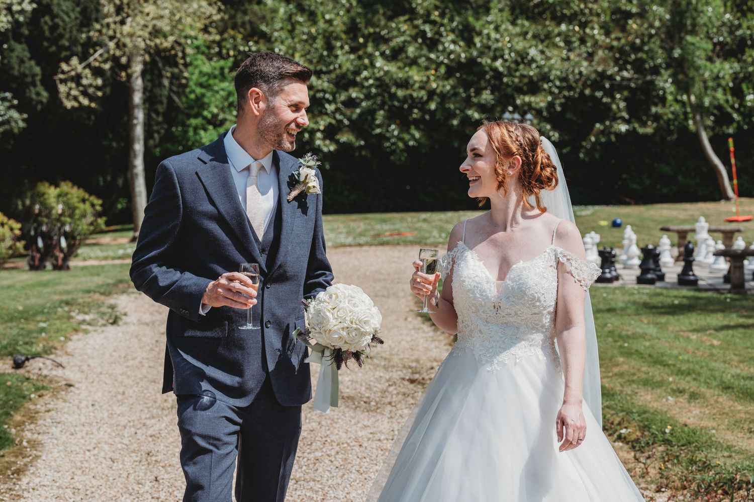 A bride and groom walk together outdoors on a garden path, with the bride holding a white bouquet.
