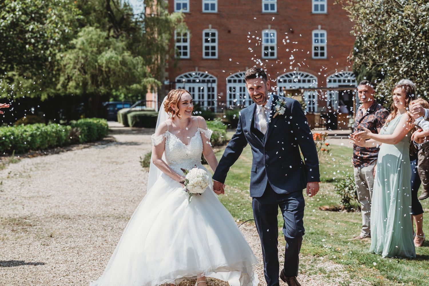 Newlyweds walk down a gravel path while guests throw confetti outside a brick building on a sunny day.