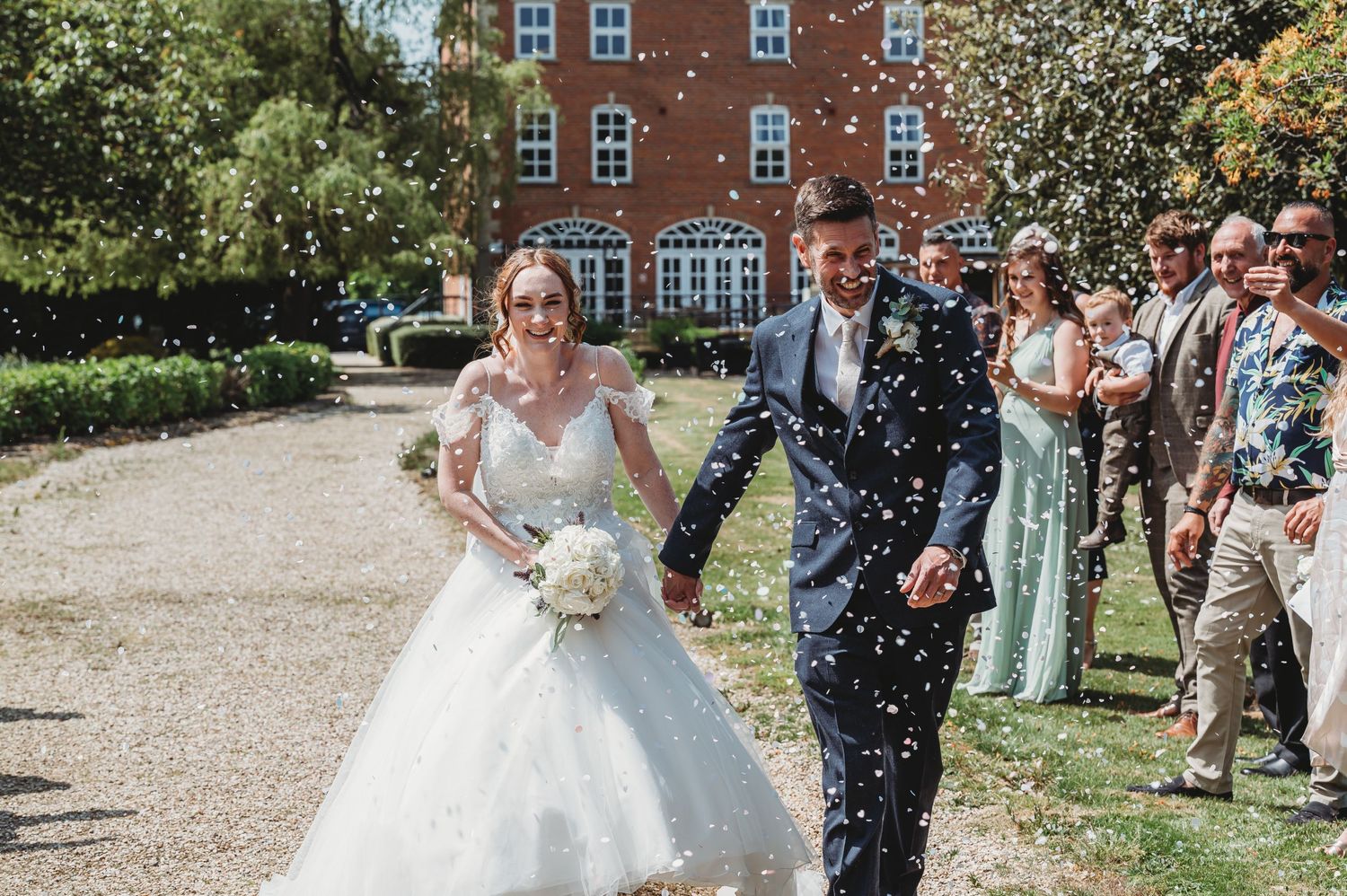 Wedding celebration at a brick mansion with guests throwing confetti along a garden path on a sunny day.