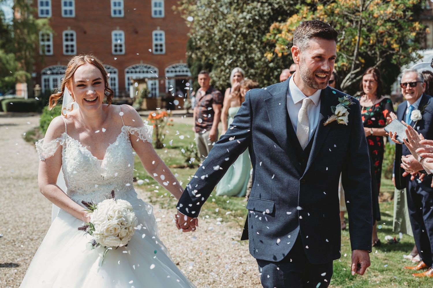 A joyful couple walks hand in hand showered in confetti surrounded by guests outside a red brick building on their wedding day.