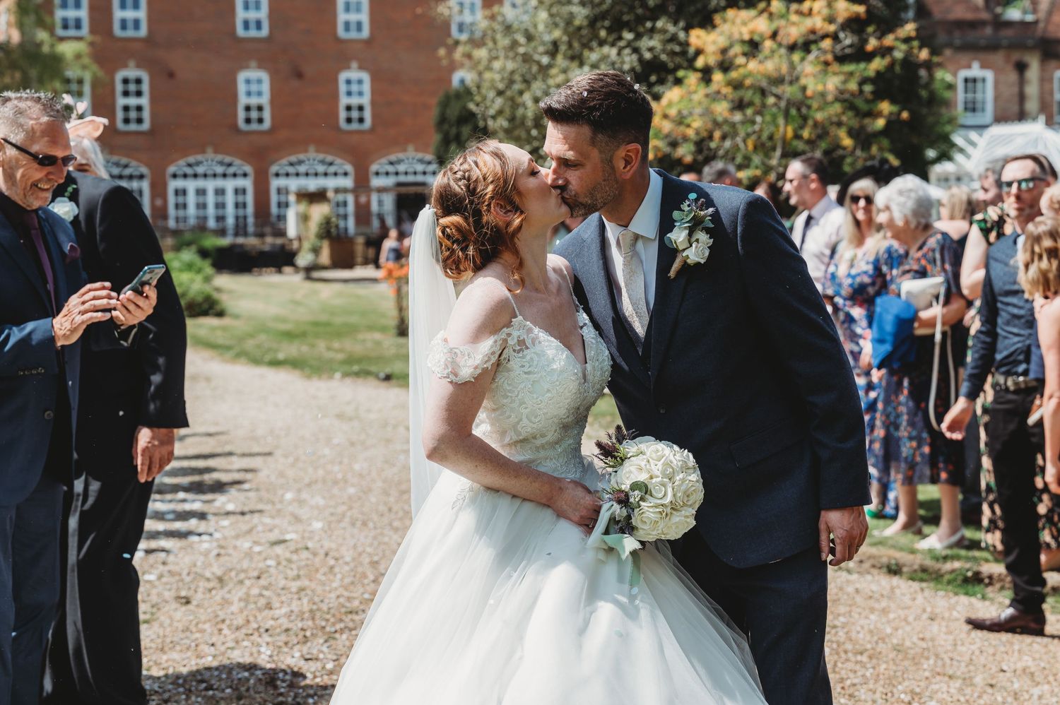 A happy wedding couple share an intimate moment on a path while guests look on during an outdoor ceremony.