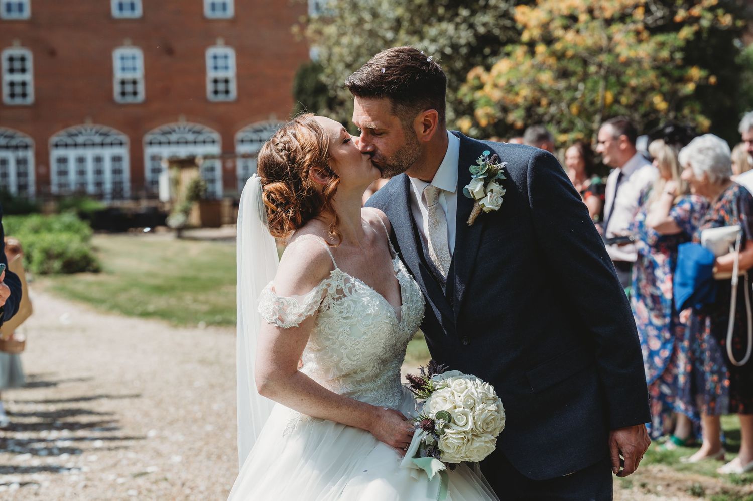 Wedding couple shares a romantic kiss on a gravel path with their guests in the background.
