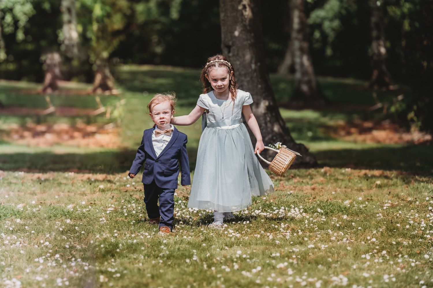 Two young children in formal attire run playfully through an autumn park during a wedding celebration.