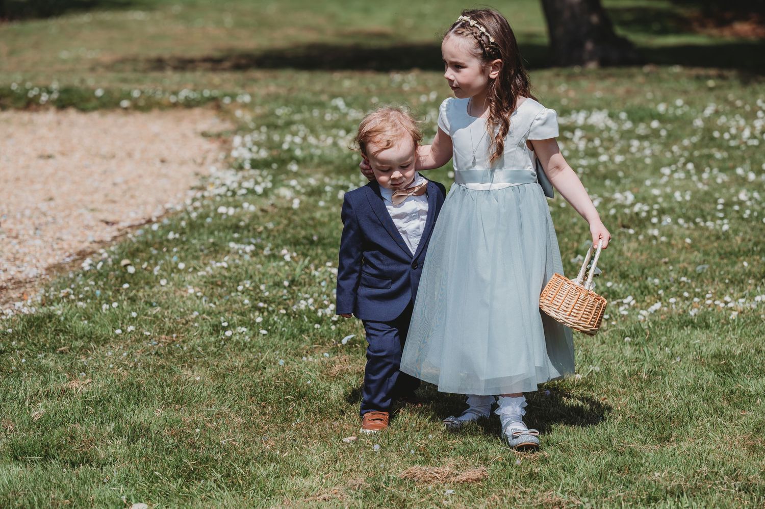 Two young children in formal attire walking together on grass with wildflowers, one carrying a small basket.