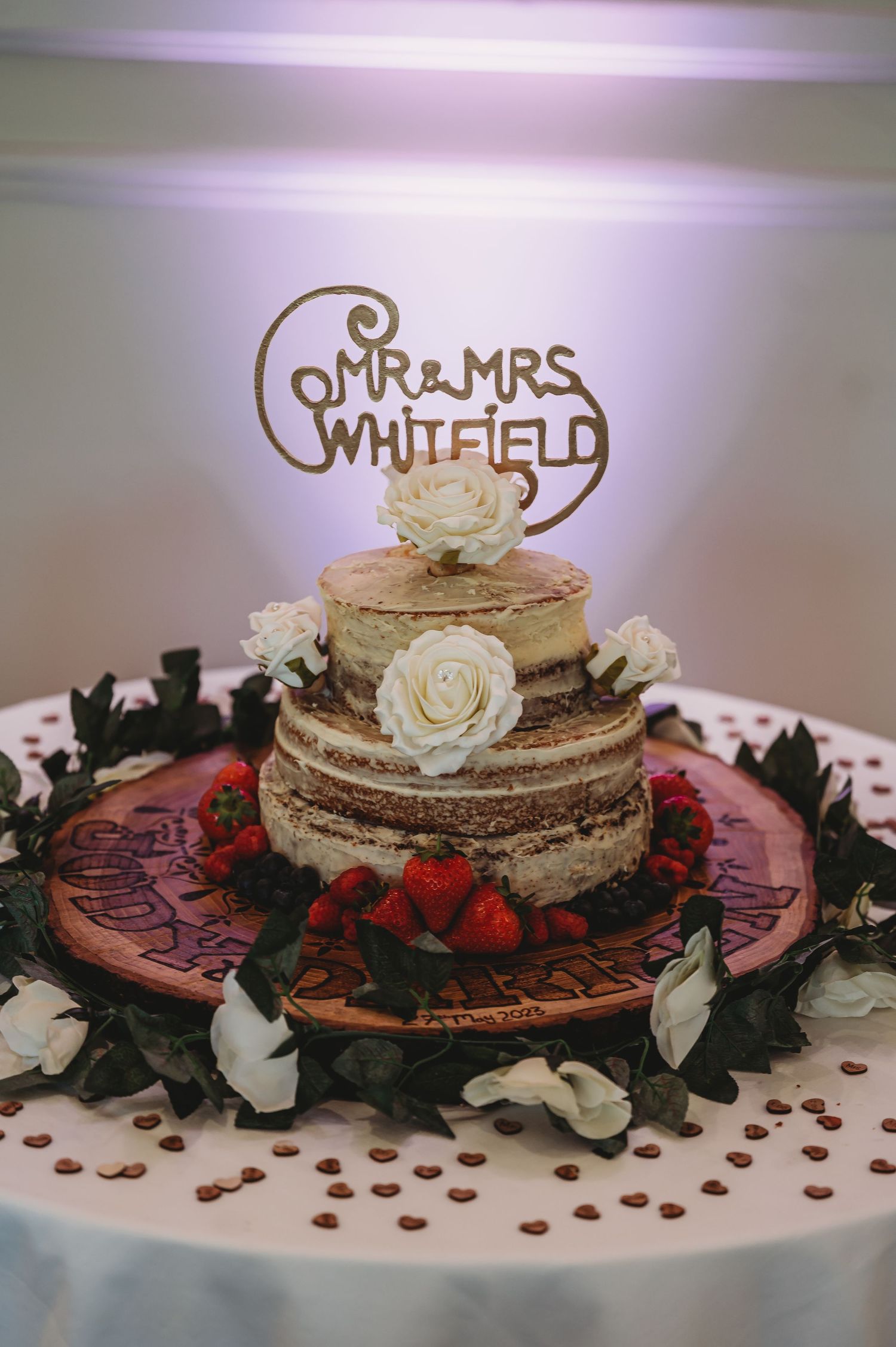 Rustic naked wedding cake decorated with white roses and strawberries features a Mr & Mrs Whitfield gold cake topper.