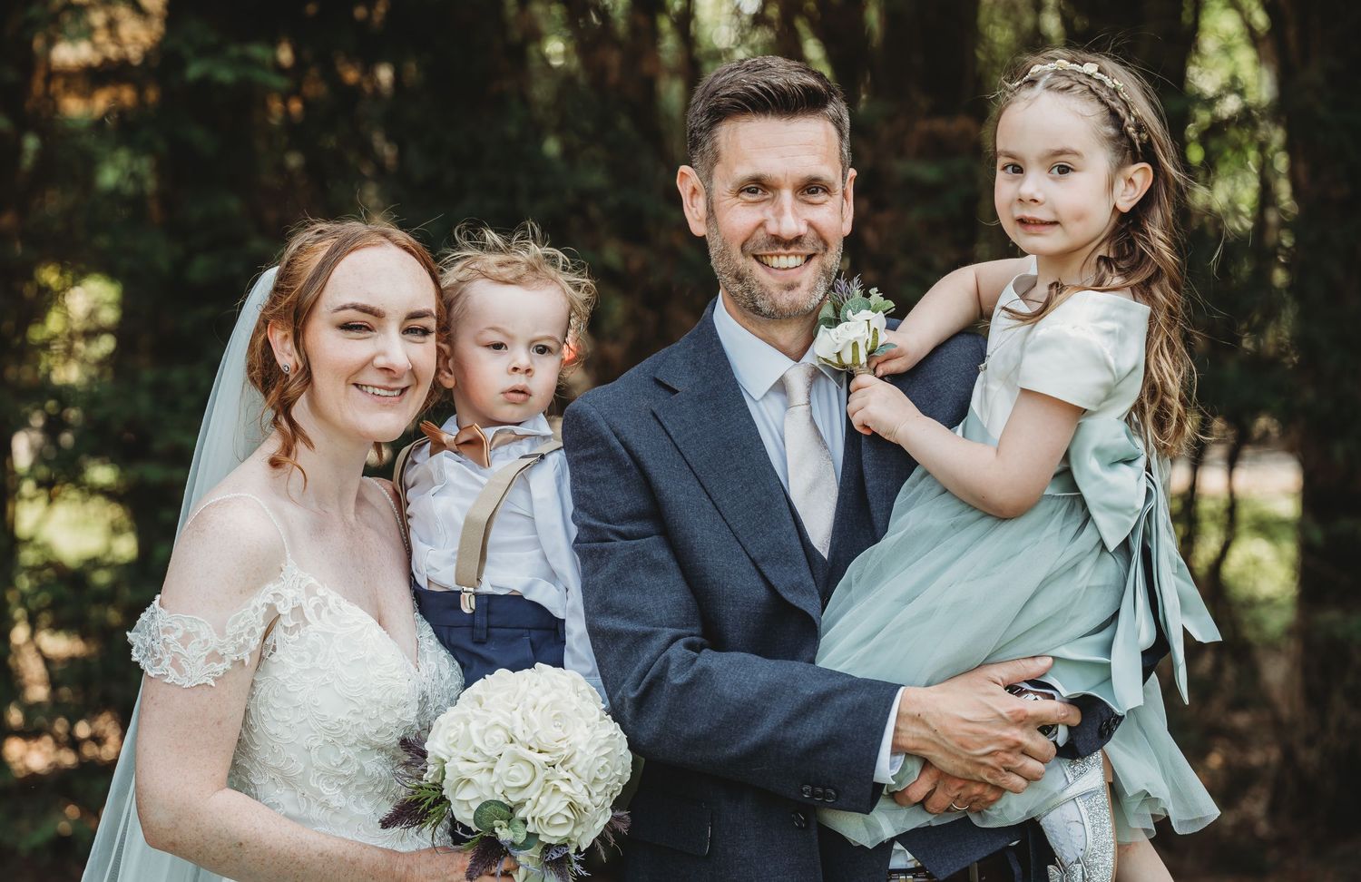 Wedding portrait of a bride and groom in formal attire with two children outdoors in a wooded setting.
