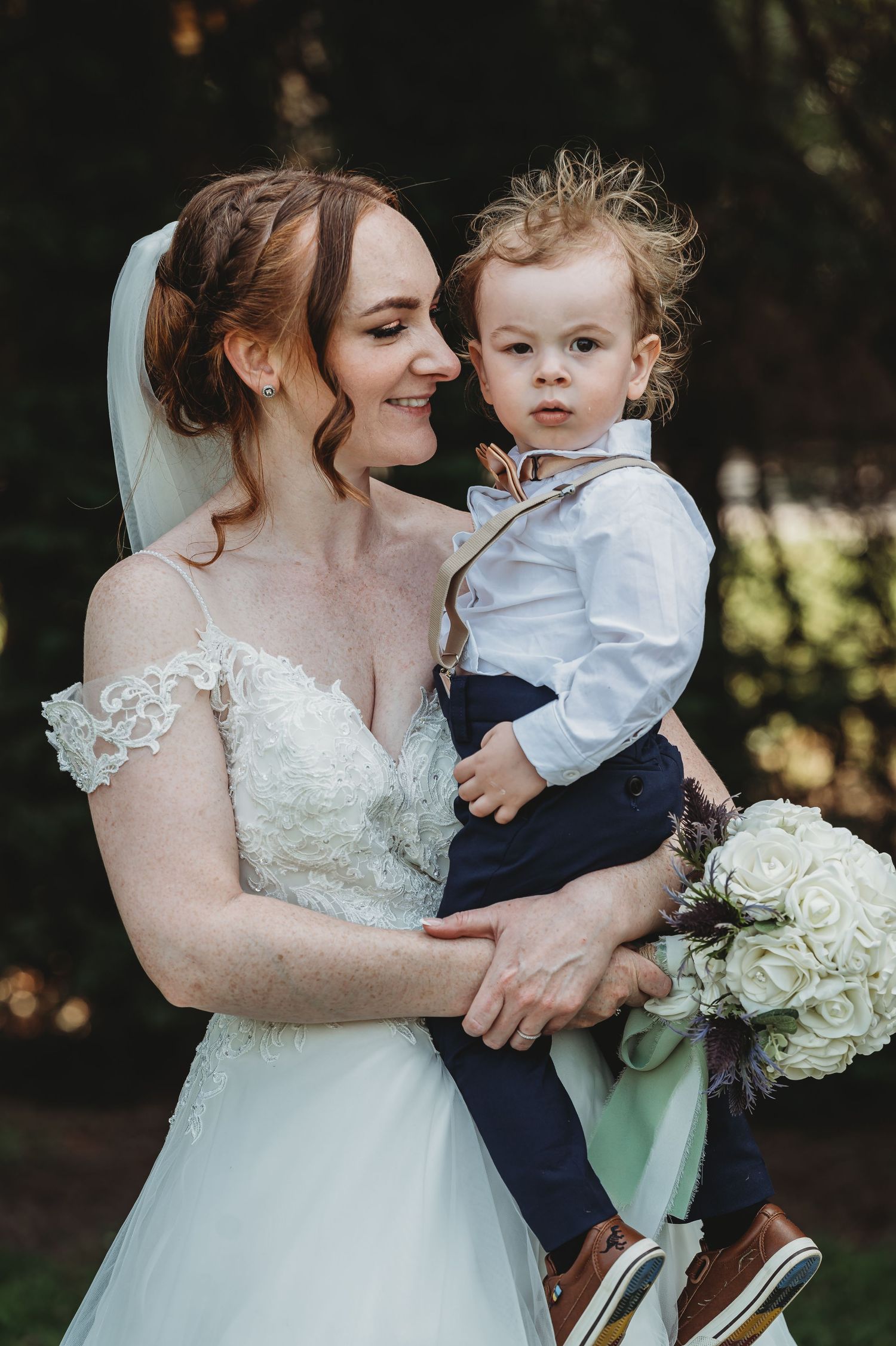 A tender moment at a wedding as a bride in a lacy white gown holds a small child in formal attire against a natural backdrop.