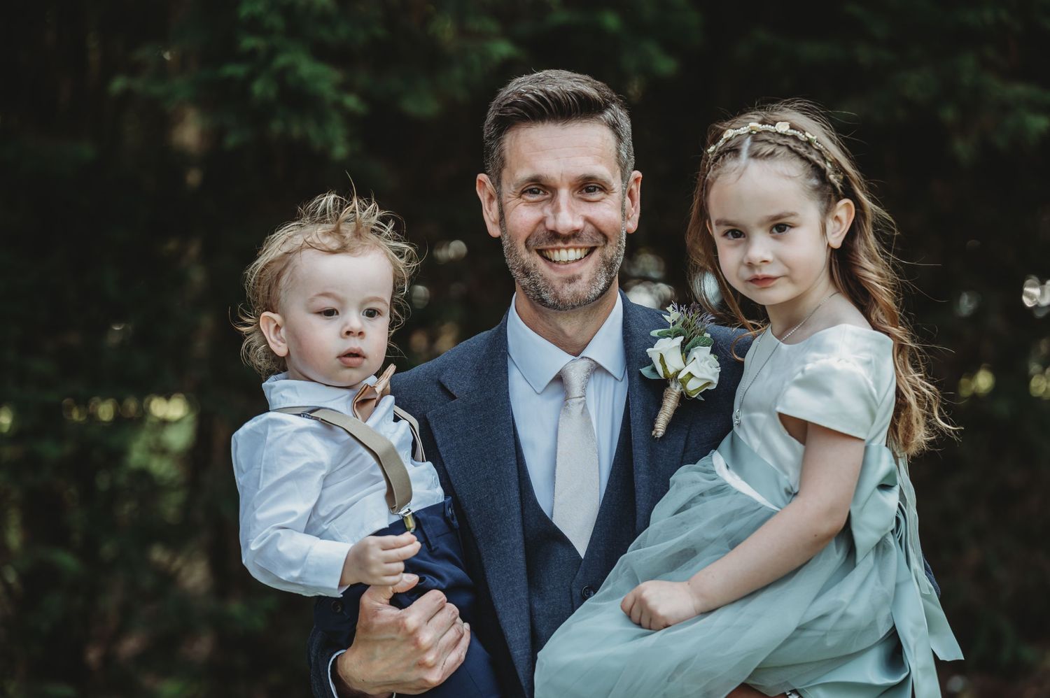 Wedding photo of well-dressed parent with two young children in formal attire outdoors against greenery.