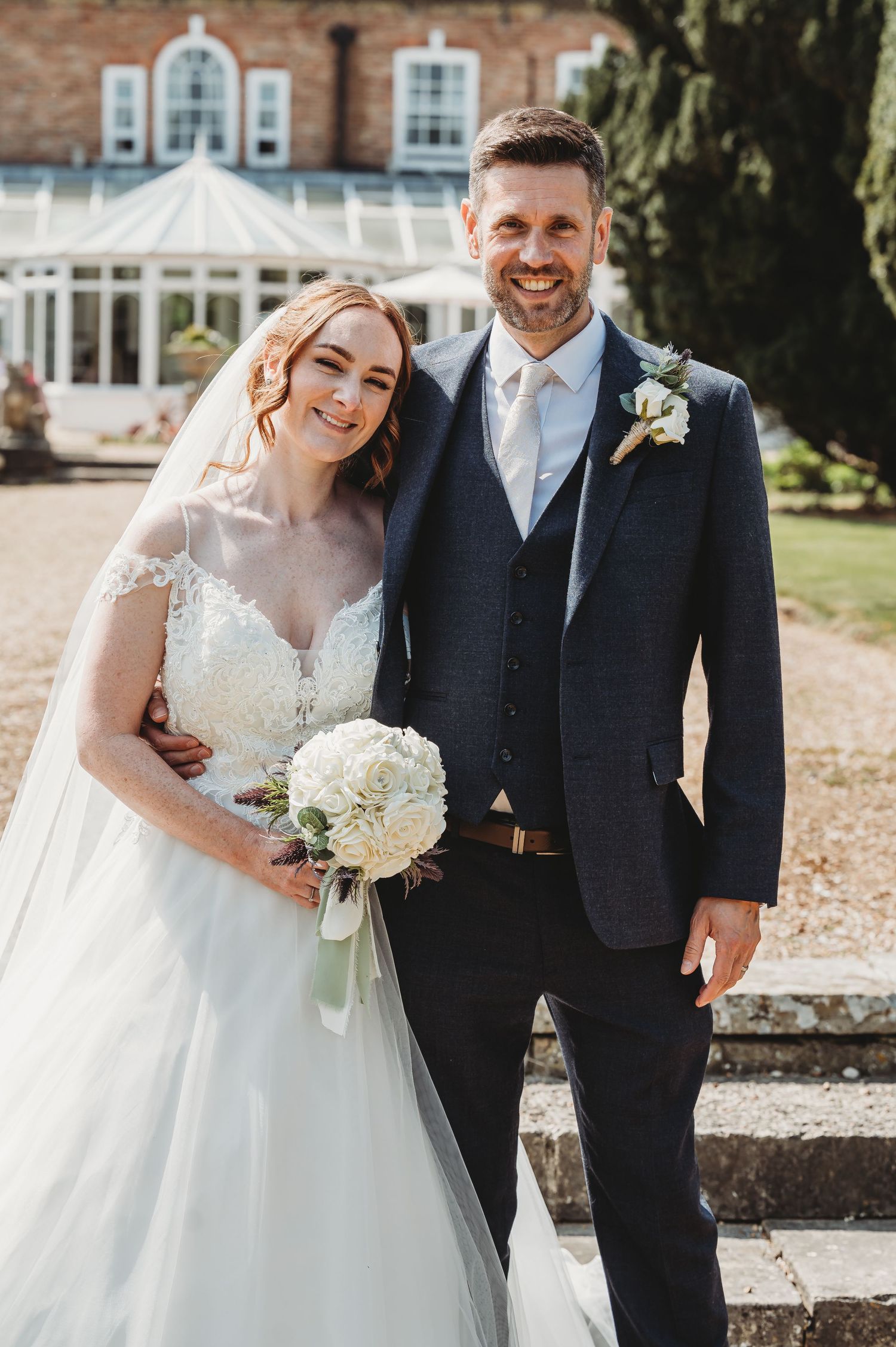 A wedding couple poses together outside a brick building with white windows and a conservatory.