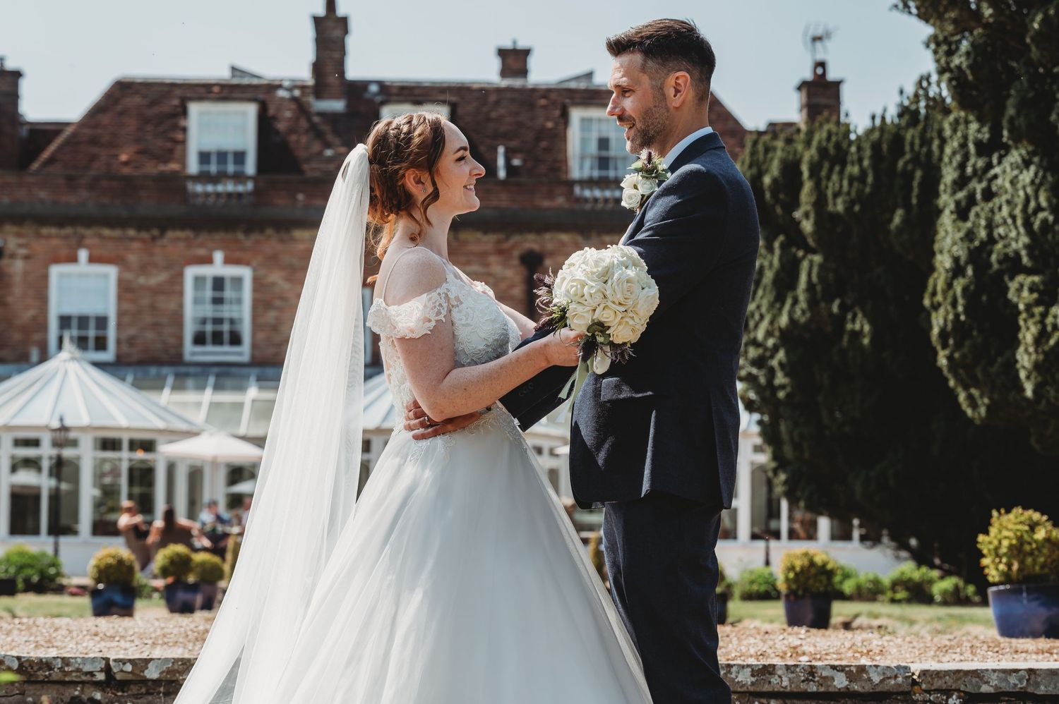 A wedding couple stands together in a garden setting with a beautiful historic building in the background.
