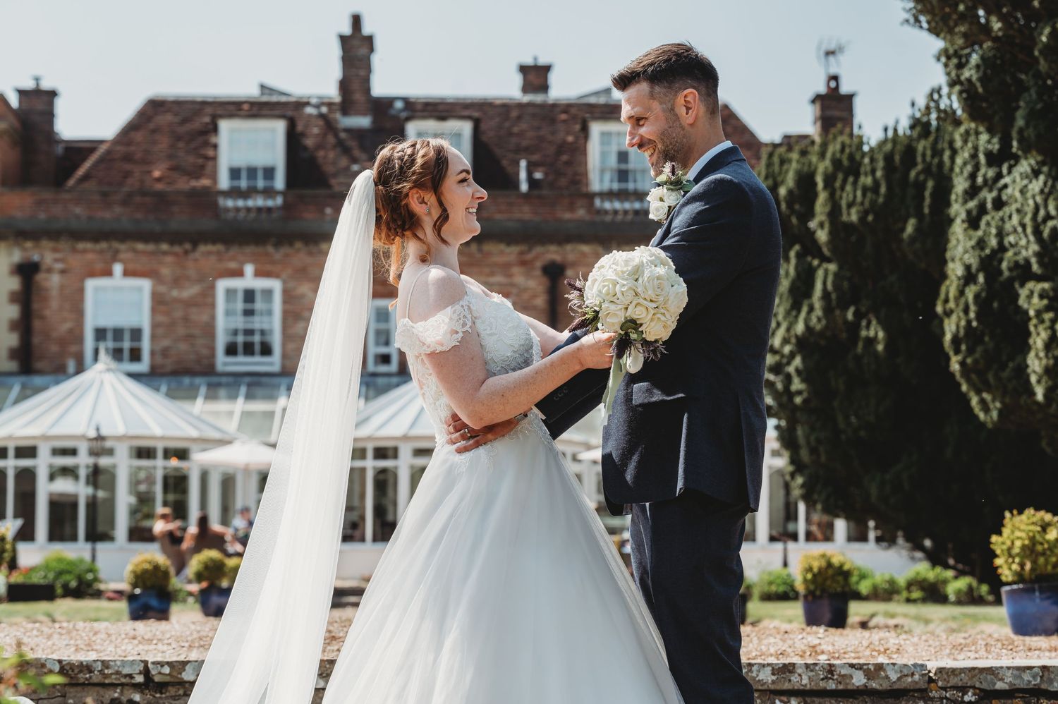 A lovely wedding ceremony takes place on a sunny day in front of a grand country house with white facade and gardens.