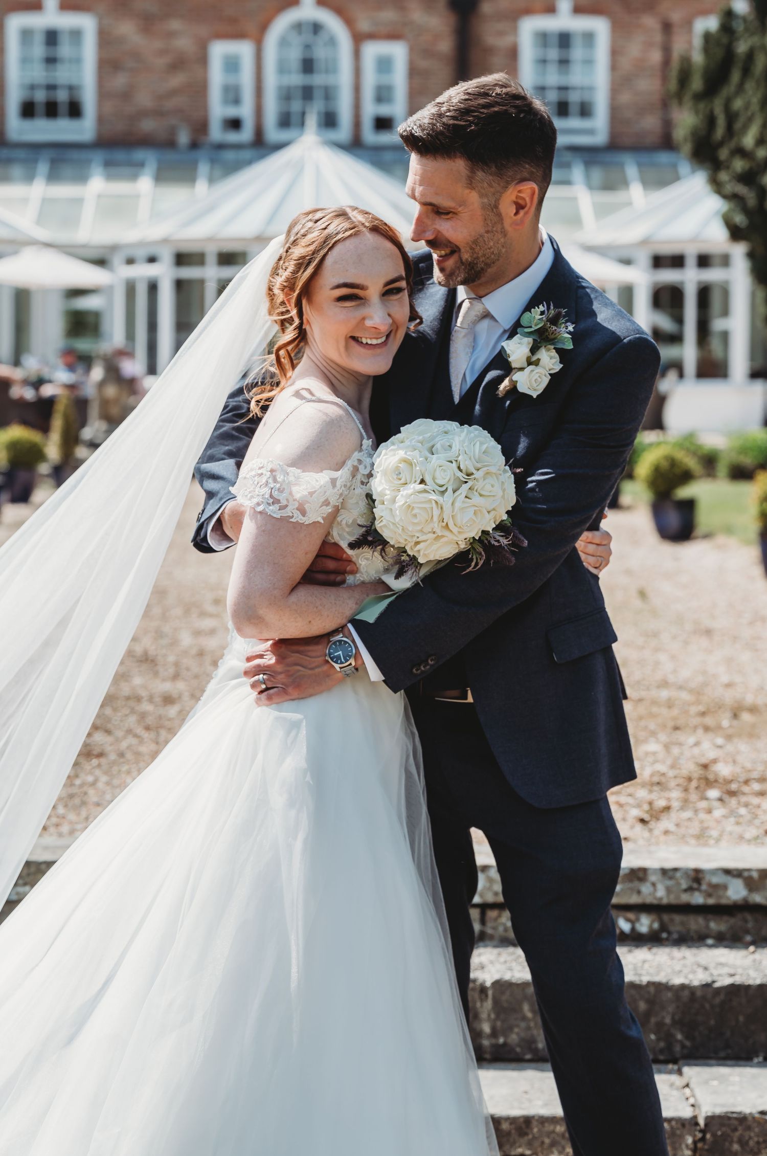 Happy wedding couple embraces outdoors in front of brick building with bride holding white floral bouquet.