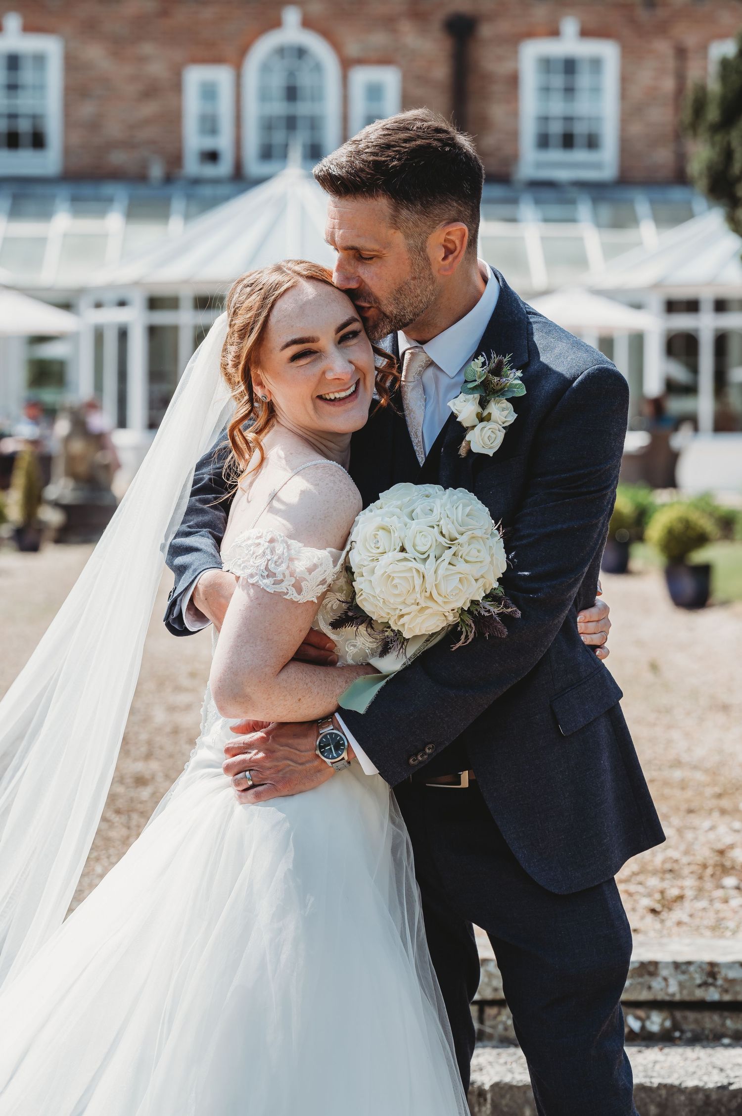 A joyful couple embraces on their wedding day outside a brick building with white windows and a conservatory.