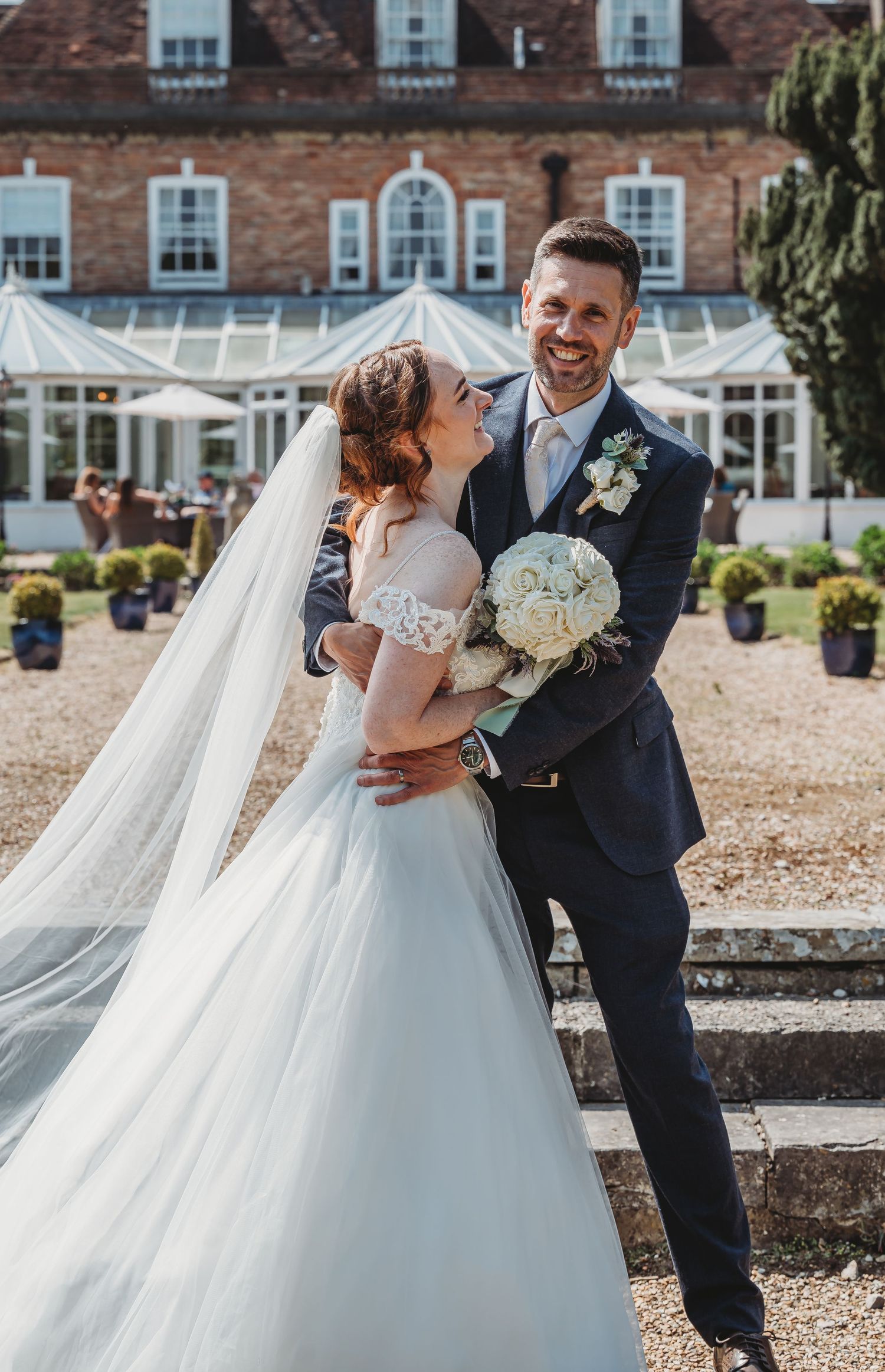 A joyful wedding couple poses in front of a beautiful brick mansion with a glass conservatory and garden.