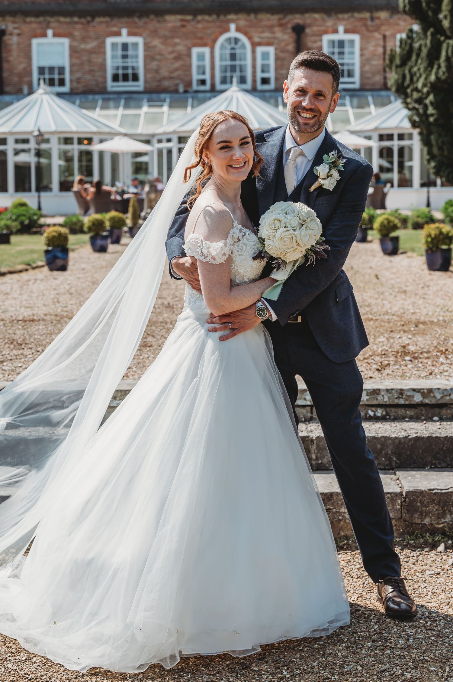 A wedding photo of a couple in formal attire posing outdoors in front of a beautiful brick mansion with white roses.