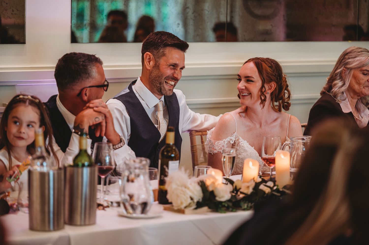 Wedding guests share laughter together at an elegant candlelit reception table decorated with white floral centerpieces.