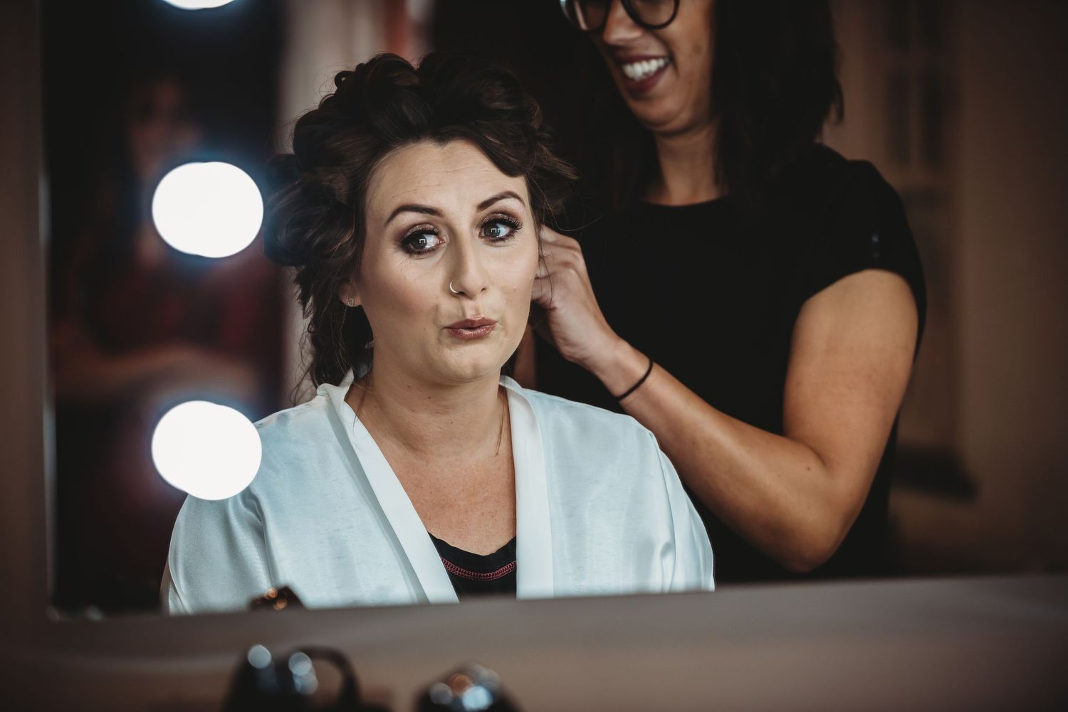 A hairstylist works on styling a client's hair in front of a mirror with bokeh lighting in the background.
