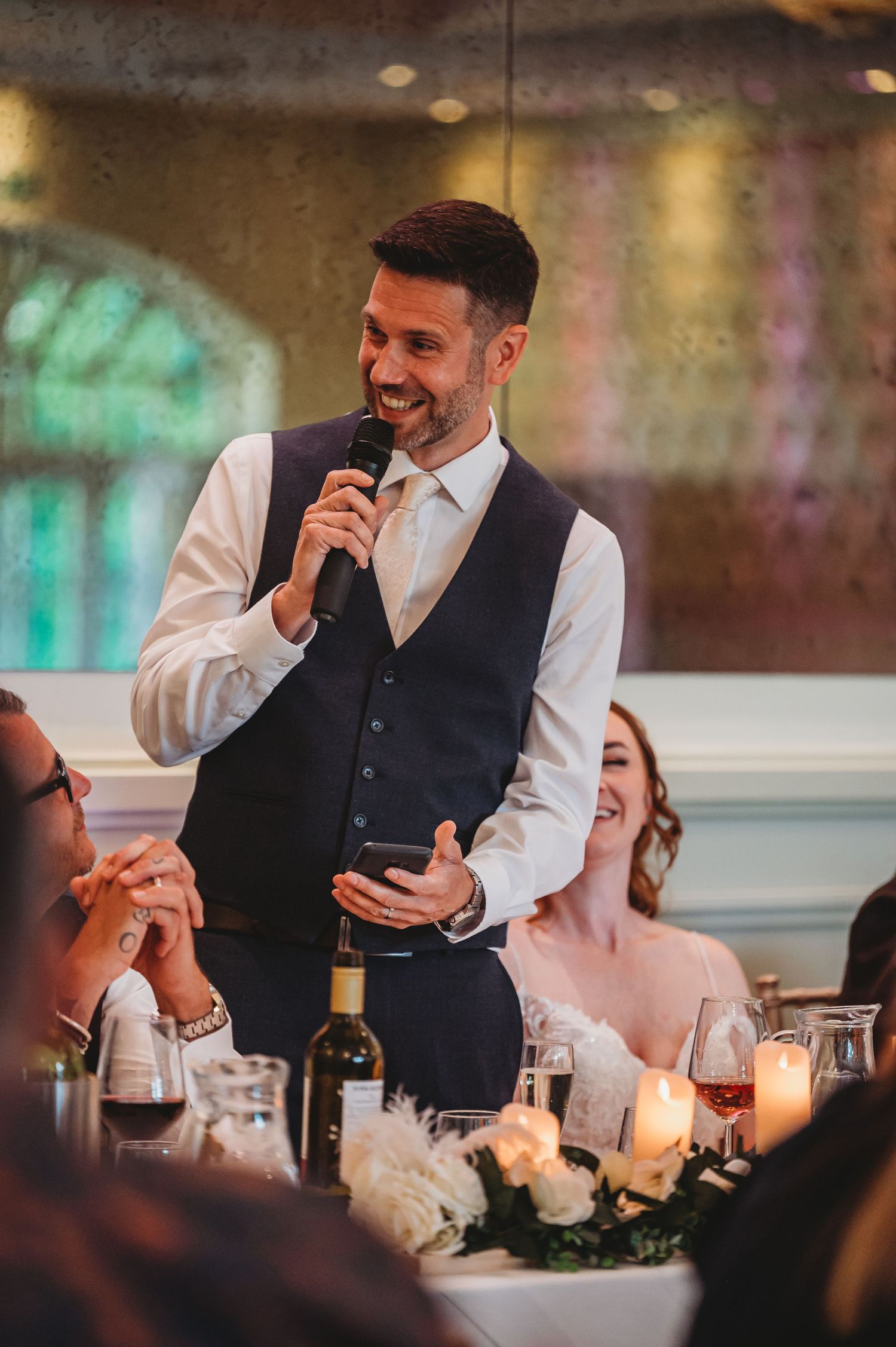 A person in a waistcoat and white shirt speaks into a microphone during a celebratory dinner reception with candlelight.