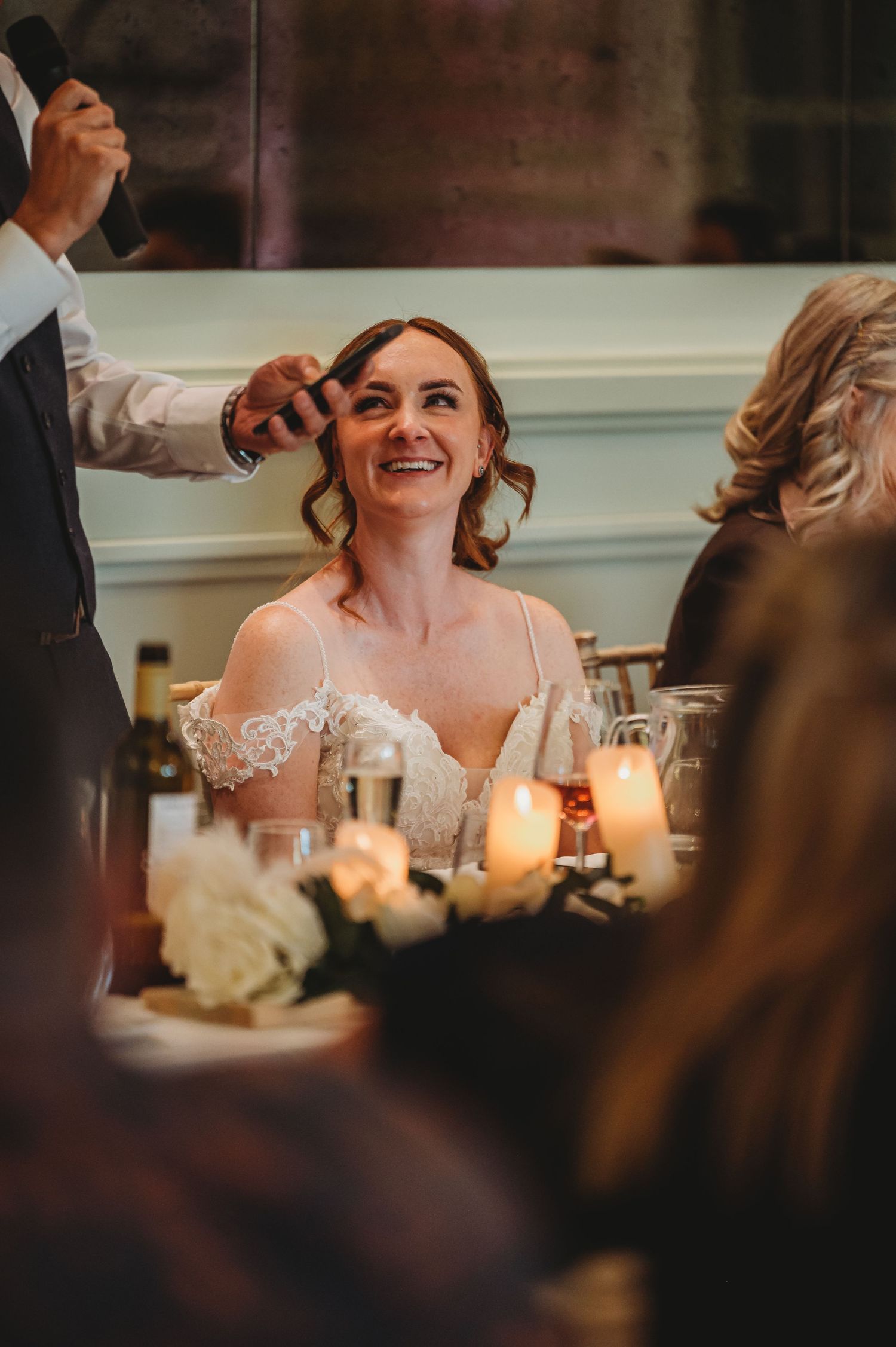 A bride in a lace off-shoulder dress laughs joyfully at her candlelit wedding reception while surrounded by guests.