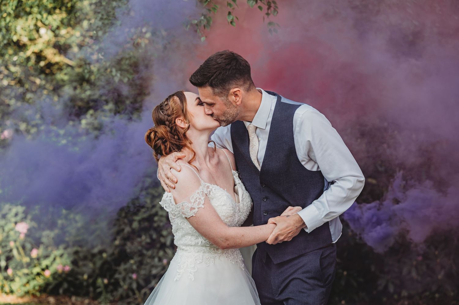 A romantic wedding photo with colorful purple and pink smoke effects in a dreamy outdoor garden setting.