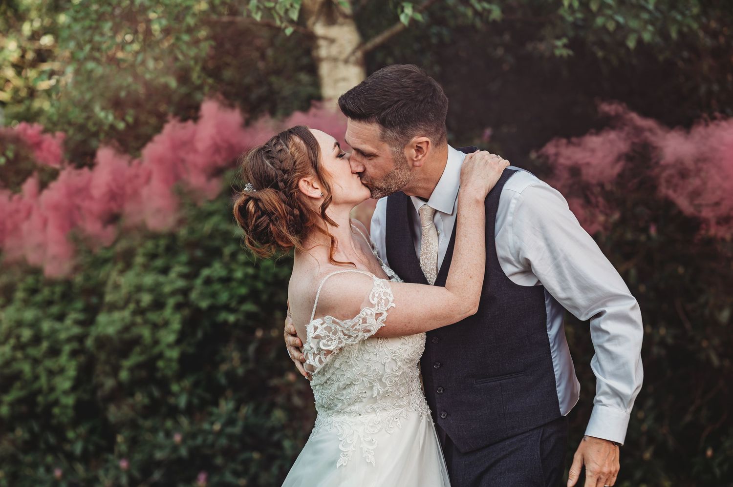 Couple shares romantic kiss in garden with pink smoke effect while wearing elegant wedding attire.
