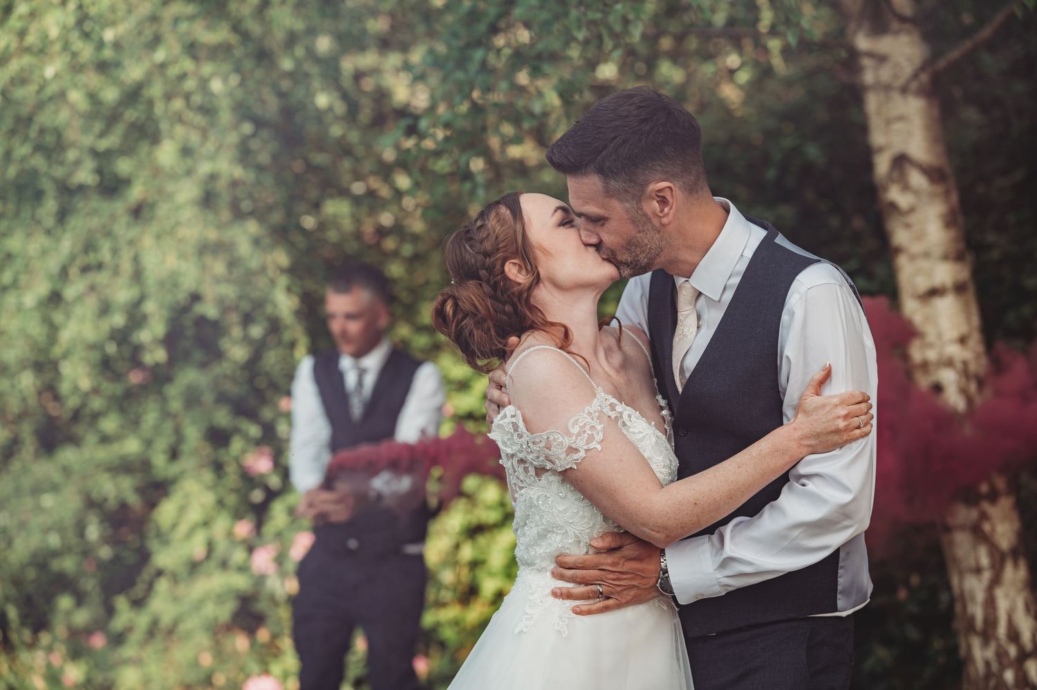 Romantic outdoor wedding scene with couple embracing in front of lush trees while groomsman stands in background.