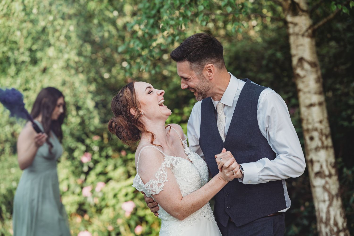 Couple dancing and smiling at outdoor wedding ceremony with bridesmaid in sage green dress visible in background.